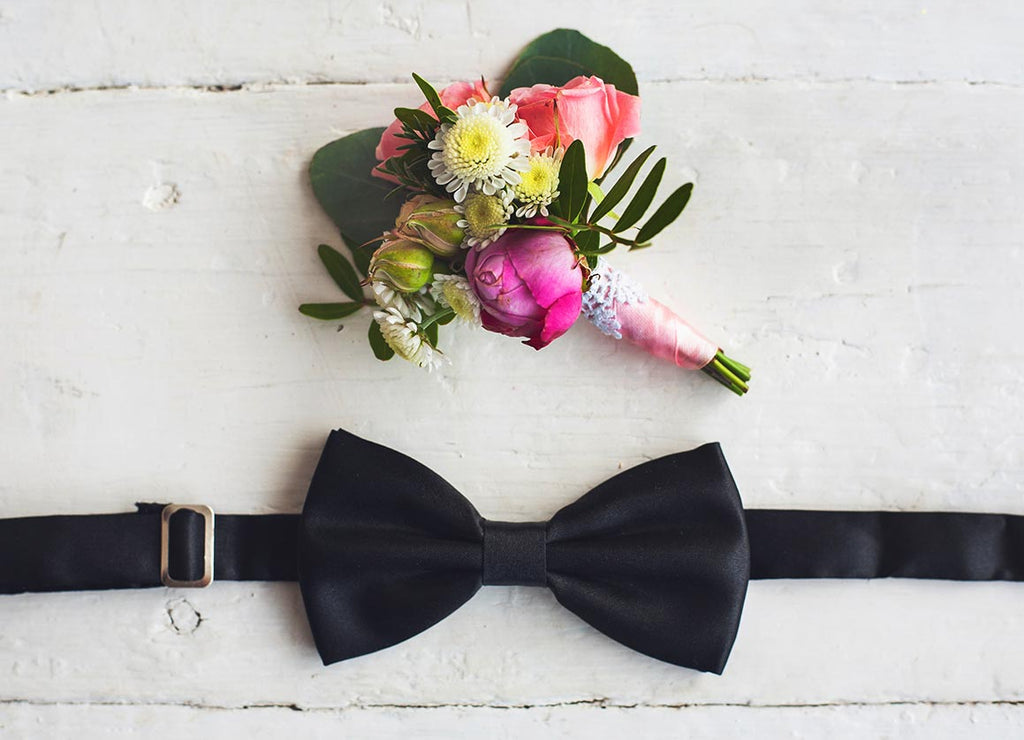Laying against a white back drop, a black bow tie sits below a colorful flower corsage.