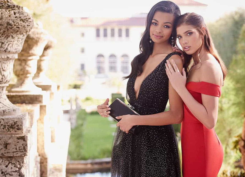 Teen girl in a red off-the-shoulder prom dress leaning on her friend in a black v-neck prom dress and holding a black clutch.