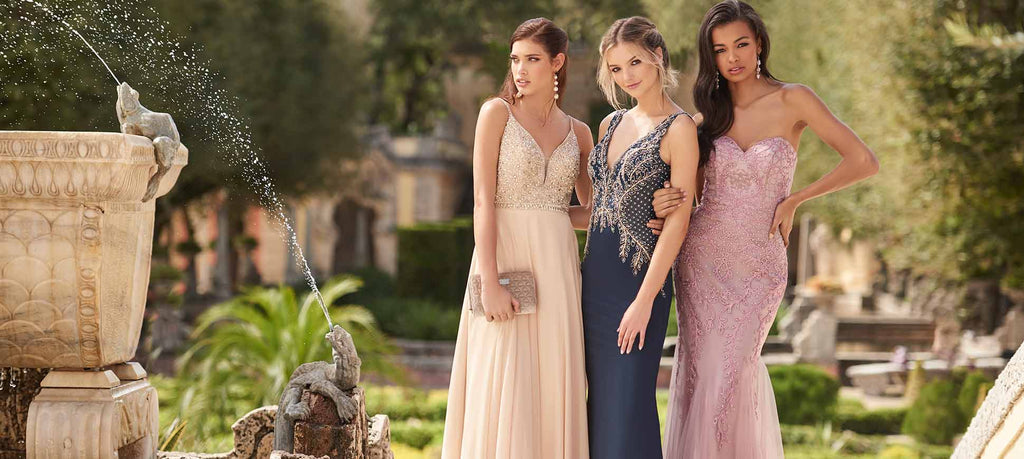 Next to a garden fountain, three teen girls wear different styles of prom dresses.