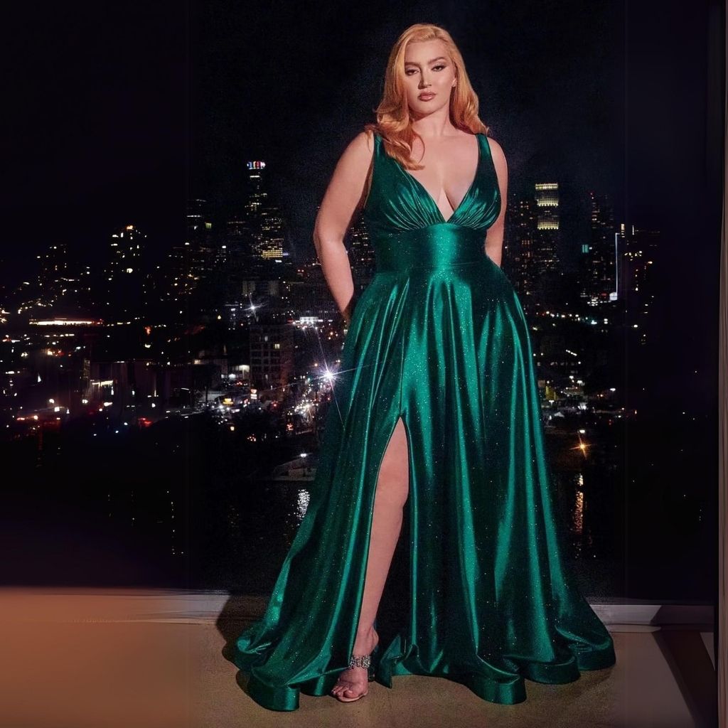 In a green plus-size empire-waist satin formal dress, a woman stands in front of a city back drop at night.
