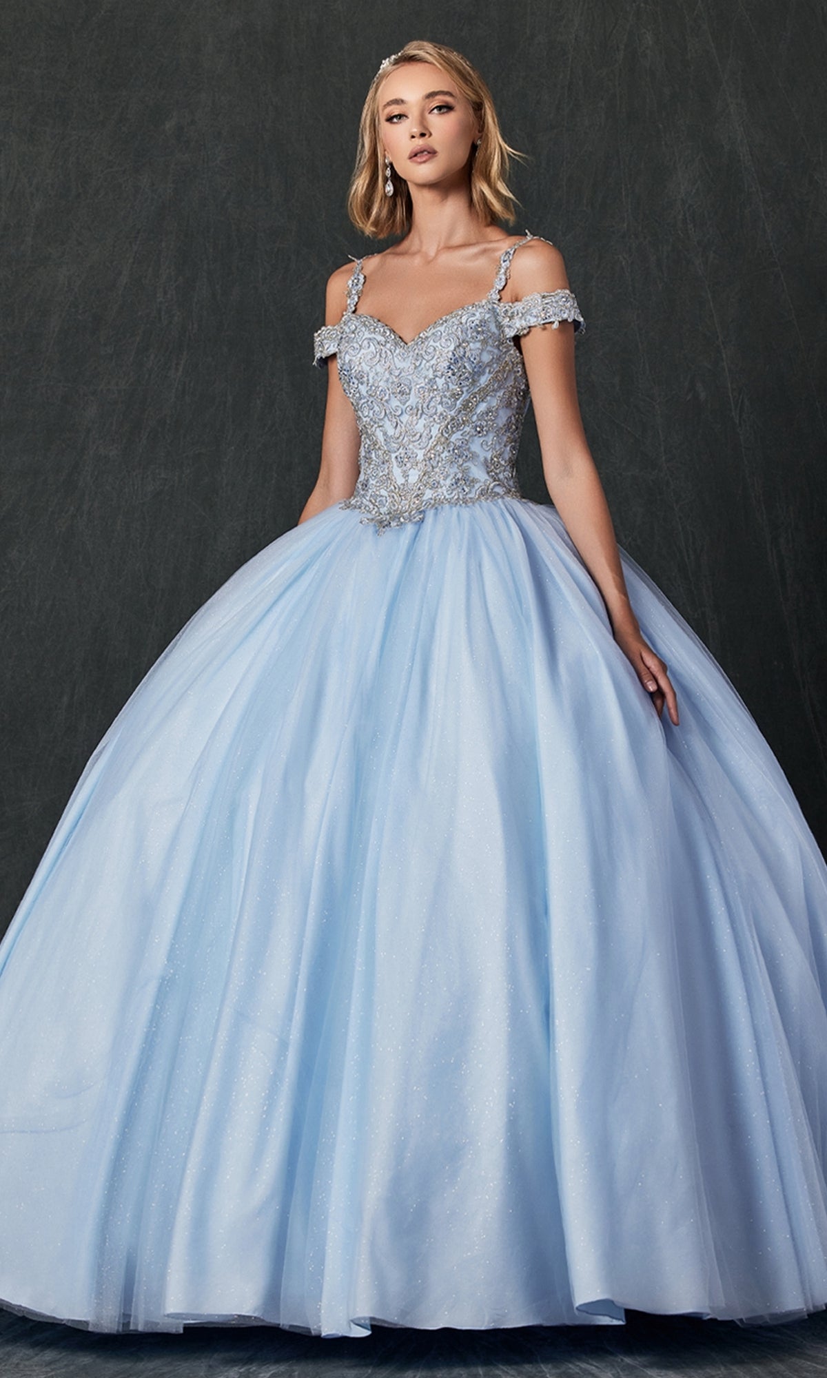 A woman models the Juliet 1430 Cold-Shoulder Glitter Quinceañera Dress, featuring a sparkling silver bodice and a voluminous light blue skirt, set against a dark background.