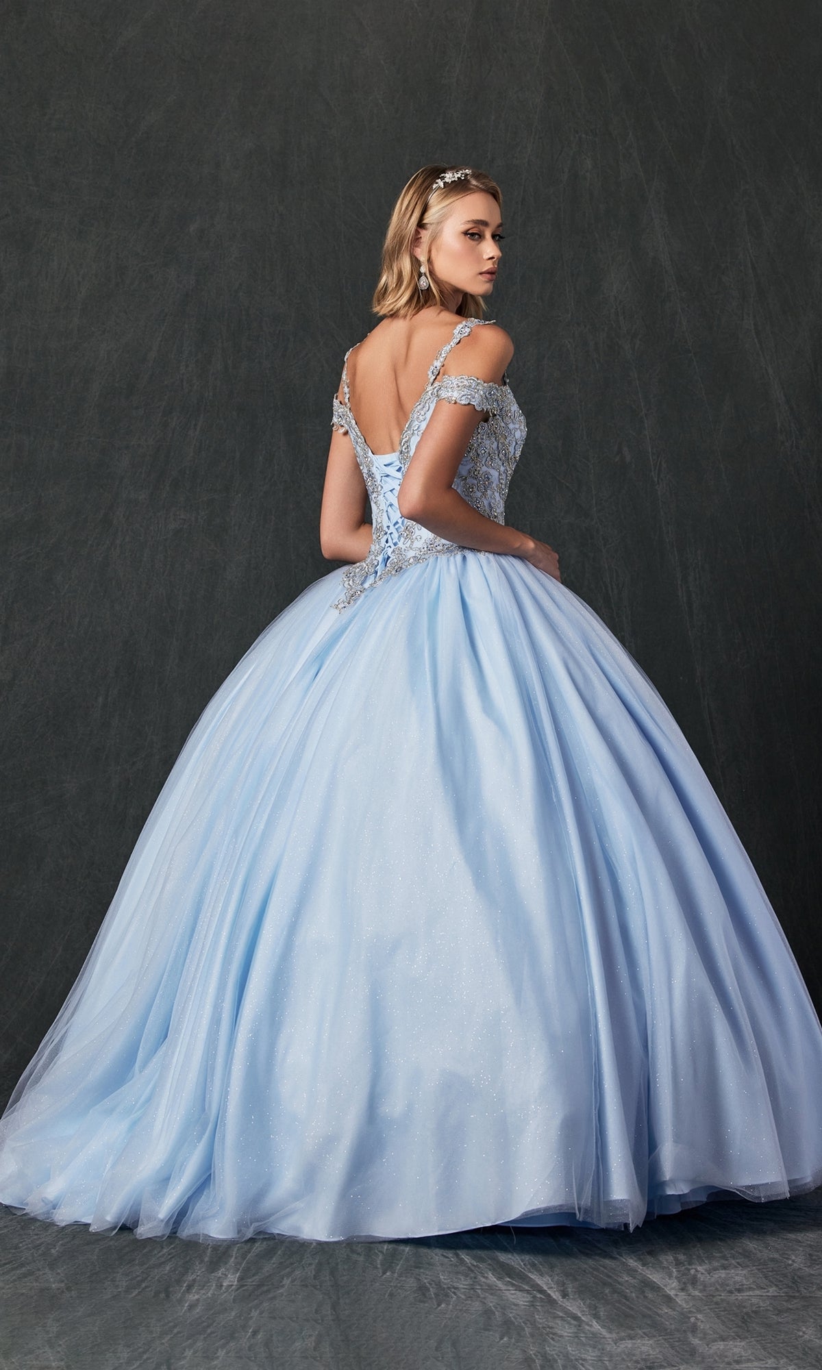 A woman wearing the Juliet 1430 Cold-Shoulder Glitter Quinceanera Dress in light blue stands against a dark textured background, gazing over her shoulder.
