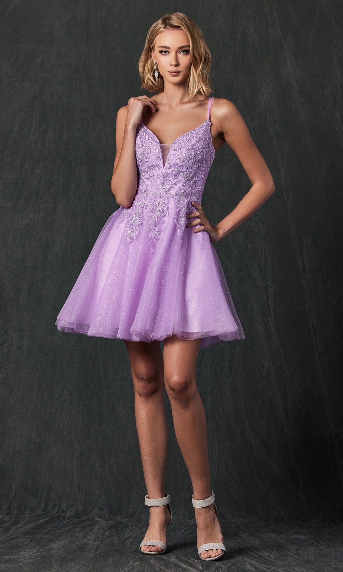 A woman wearing the Glitter-Tulle Short Babydoll Homecoming Dress 857 in lavender with lace details and spaghetti straps stands against a dark, textured background.