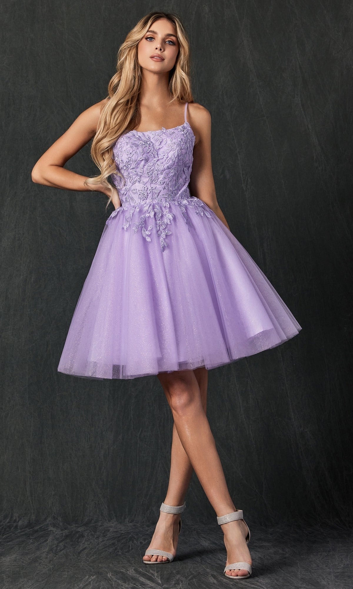 A woman models the Embroidered-Corset Babydoll Homecoming Dress 860, a sleeveless lilac dress with floral lace, embroidery, and a tulle skirt, standing against a dark textured background.