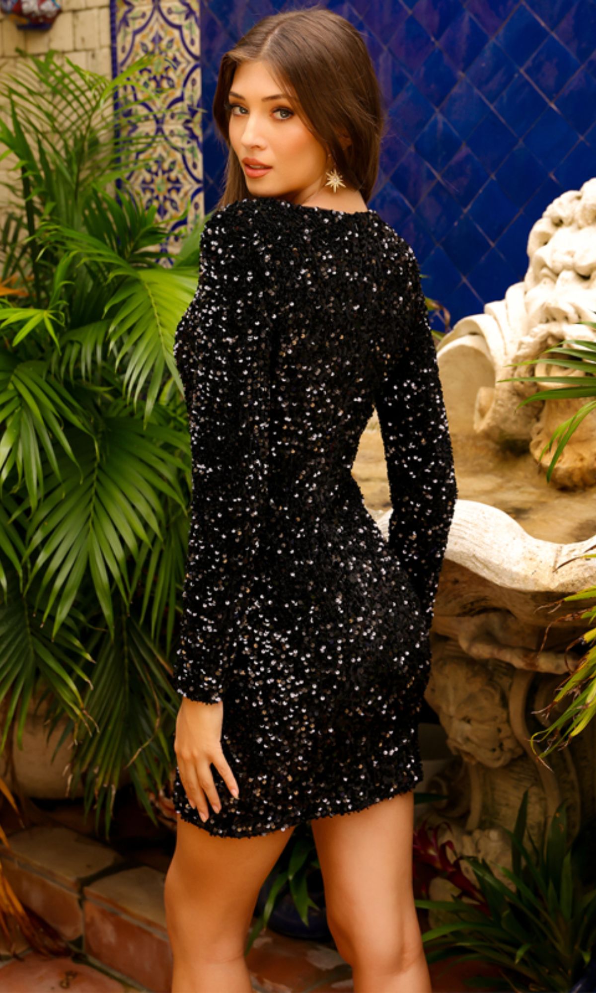 Woman standing outdoors by plants and a tiled wall, wearing the Long Sleeve Short Sequin Homecoming Dress BZ029S with black sequins, looking over her shoulder at the camera.