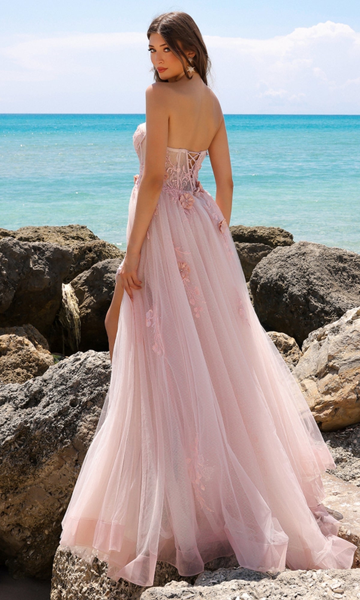 A woman in the Strapless Sheer-Corset Long Tulle Prom Dress TM1002 stands on rocks by the sea, looking over her shoulder.