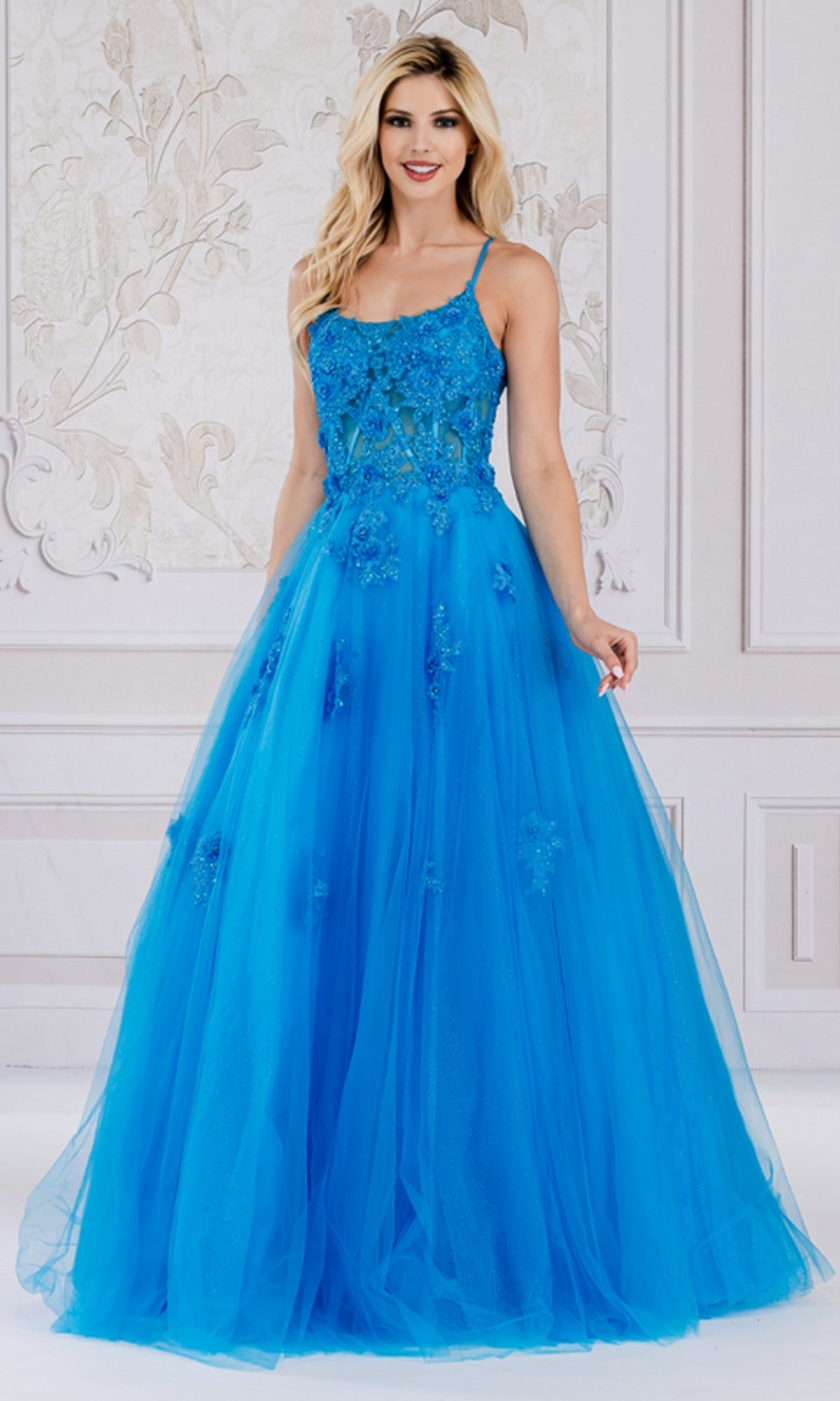 A woman stands in a decorated room wearing the Embroidered-Tulle Long A-Line Prom Ball Gown 7035, featuring a bright blue, floor-length design with floral embellishments.