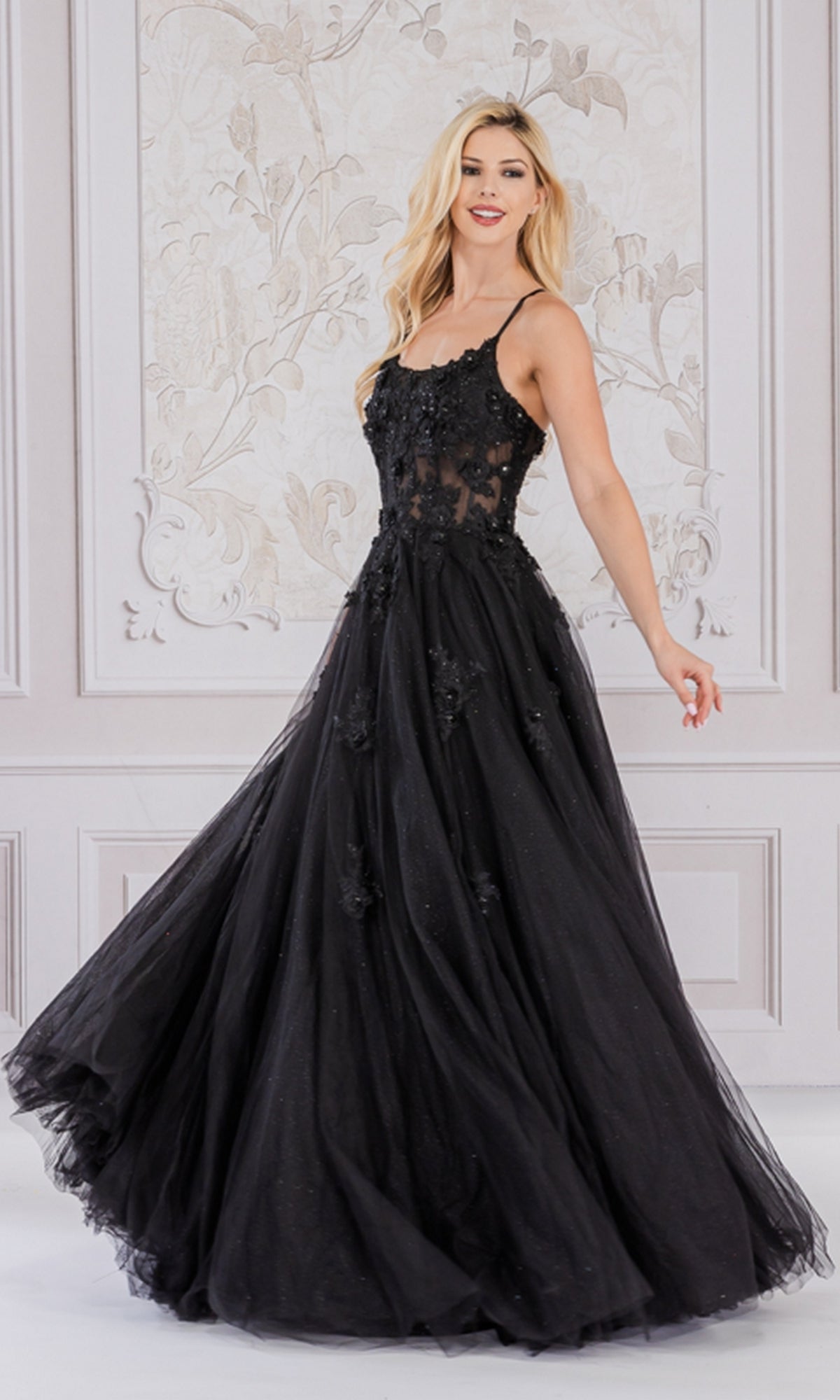 A woman twirls in a softly decorated room, showcasing the Embroidered-Tulle Long A-Line Prom Ball Gown 7035—a black, floor-length dress with floral embroidery, a sheer bodice, sleeveless design, and a full skirt.