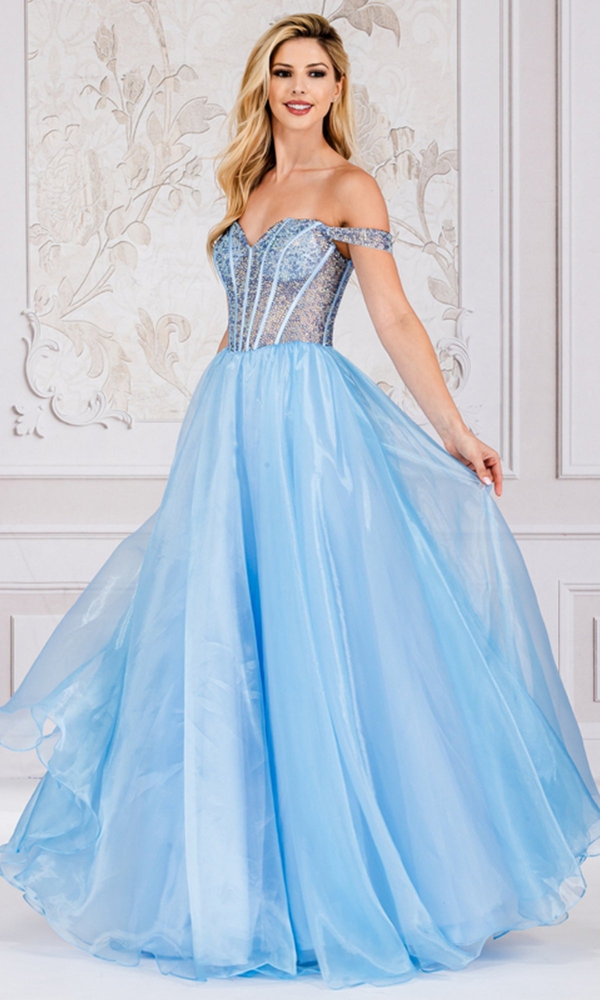 A woman stands in an elegantly decorated room wearing the Organza Off-Shoulder Bodice Prom Ball Gown 7040, featuring a light blue fitted semi-sheer bodice and a flowing organza skirt.