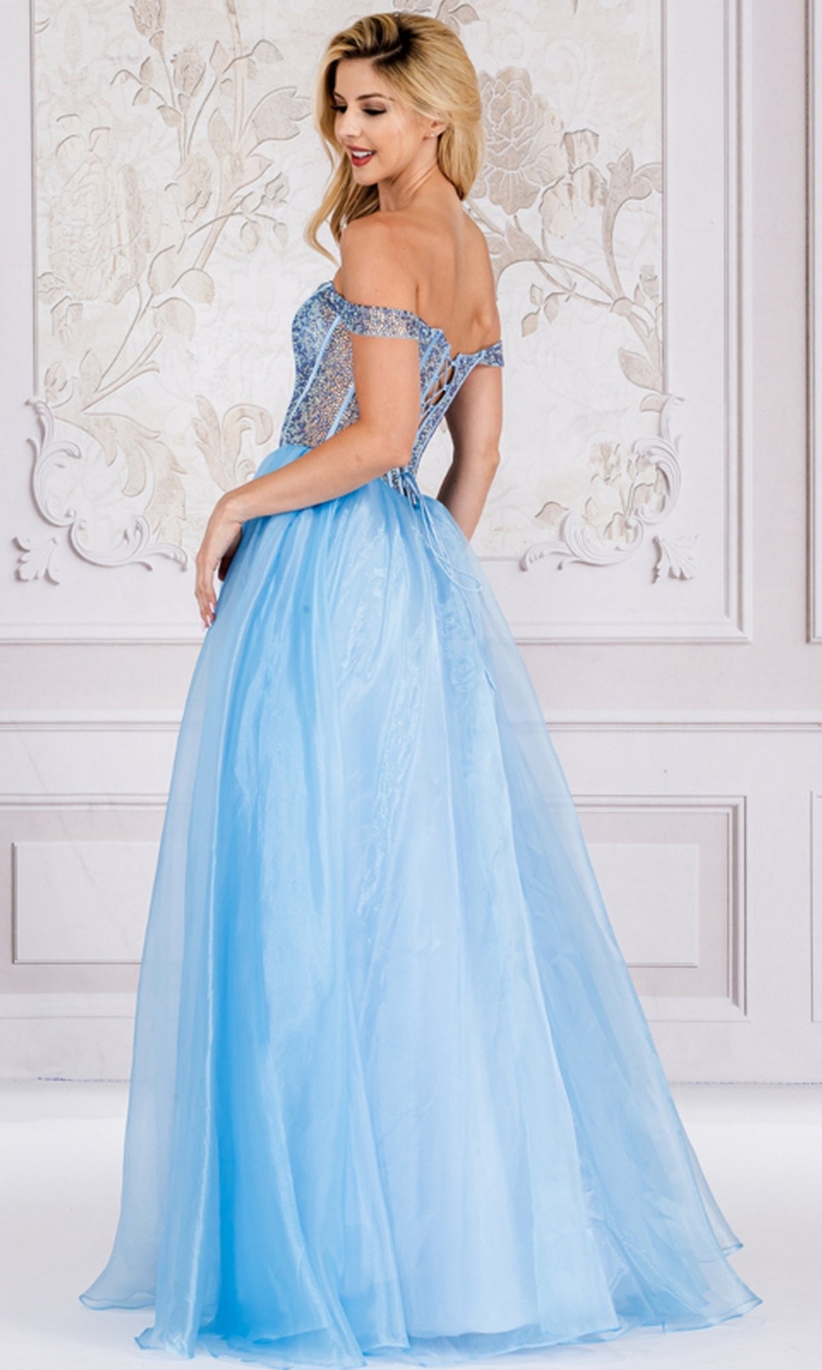 A woman stands in an ornate room wearing the Organza Off-Shoulder Bodice Prom Ball Gown 7040, which features a fitted, embellished bodice and a flowing organza skirt.