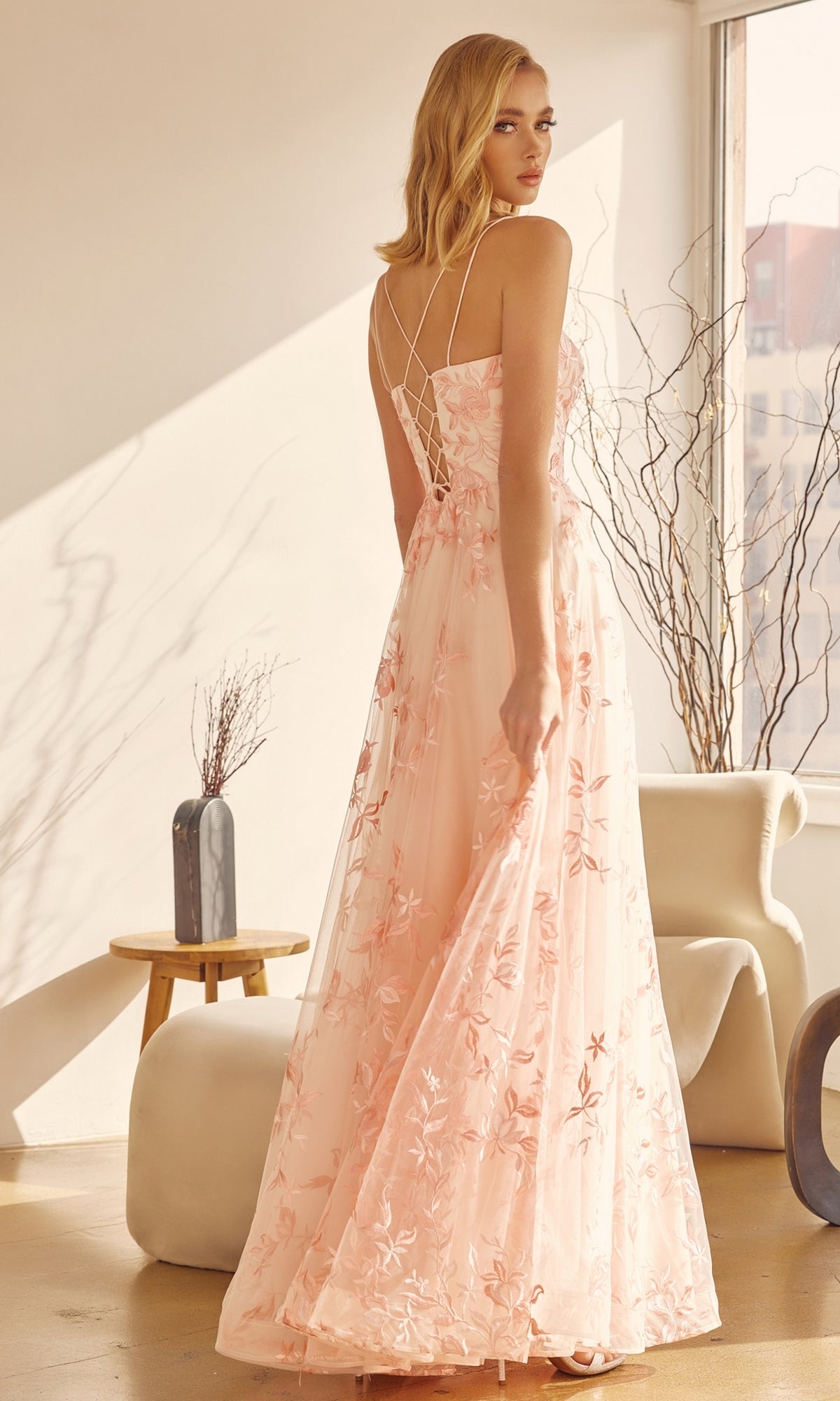 A woman stands indoors in natural light, wearing the Embroidered-Lace Long A-Line Prom Ball Gown 279 with thin straps and a lace-up back, glancing over her shoulder amid modern decor.