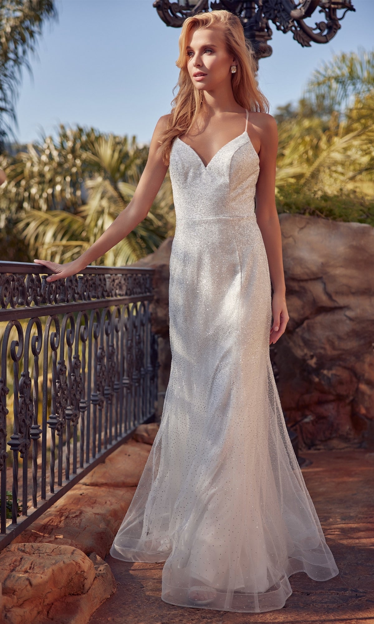 A woman in the Glitter-Mesh Long White Mermaid Formal Dress 271W with thin straps stands outdoors by a wrought iron railing, surrounded by trees and rocks.