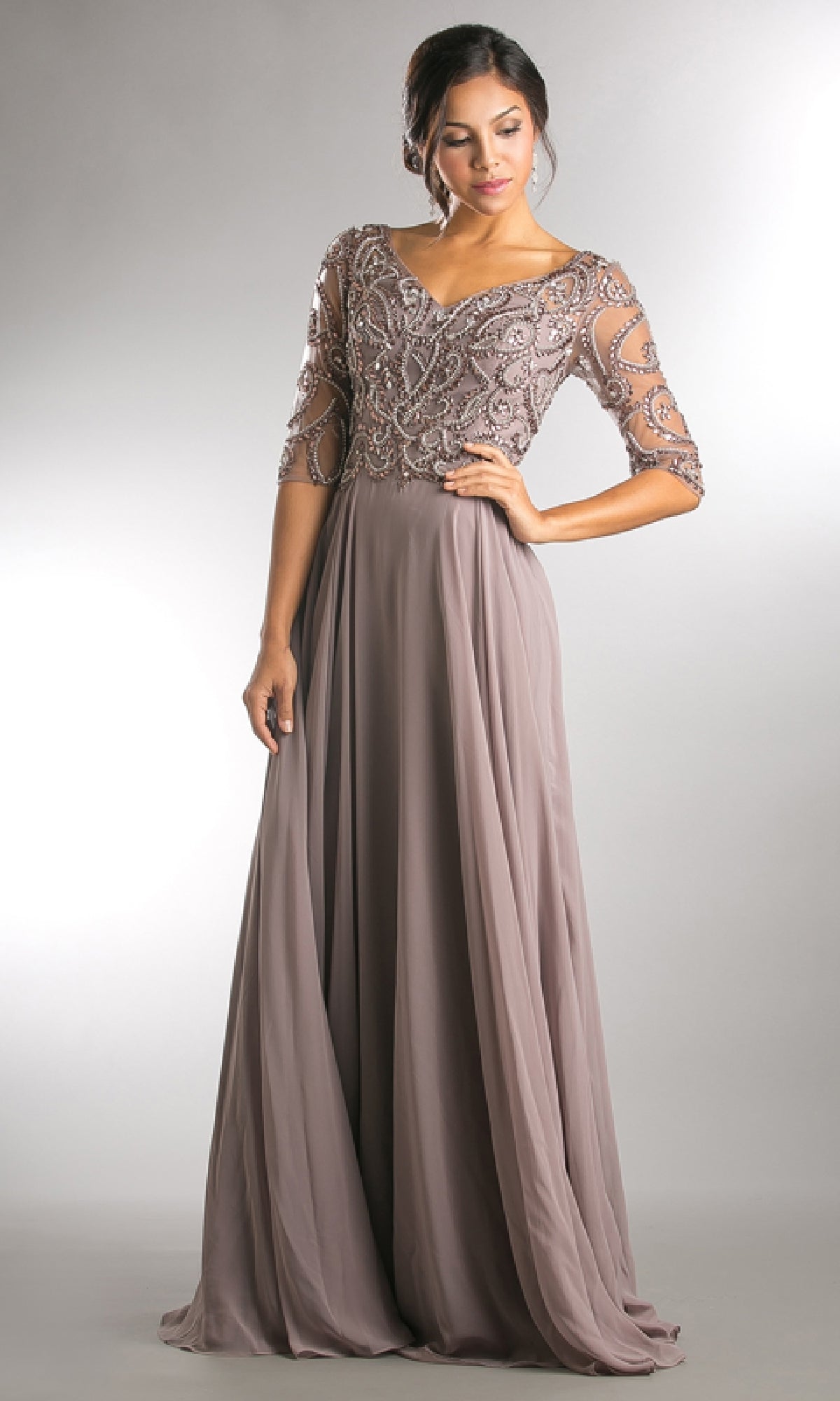 A woman poses in the Amelia Couture 7046 sleeved long formal dress, featuring beaded embroidery on the bodice and sheer half sleeves, set against a plain studio background.
