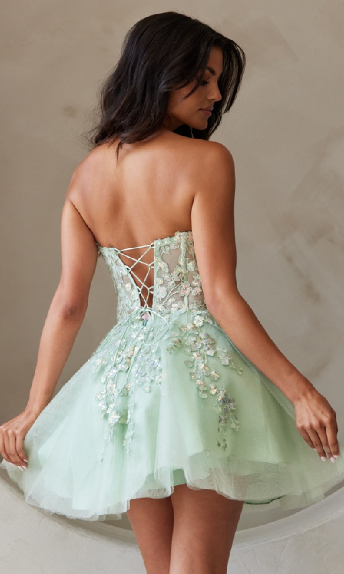 A woman models the Strapless Short Pastel Homecoming Dress BZ9024S by Amelia Couture in light green, featuring floral embroidery, a lace-up back, and tulle skirt, set against a neutral background.