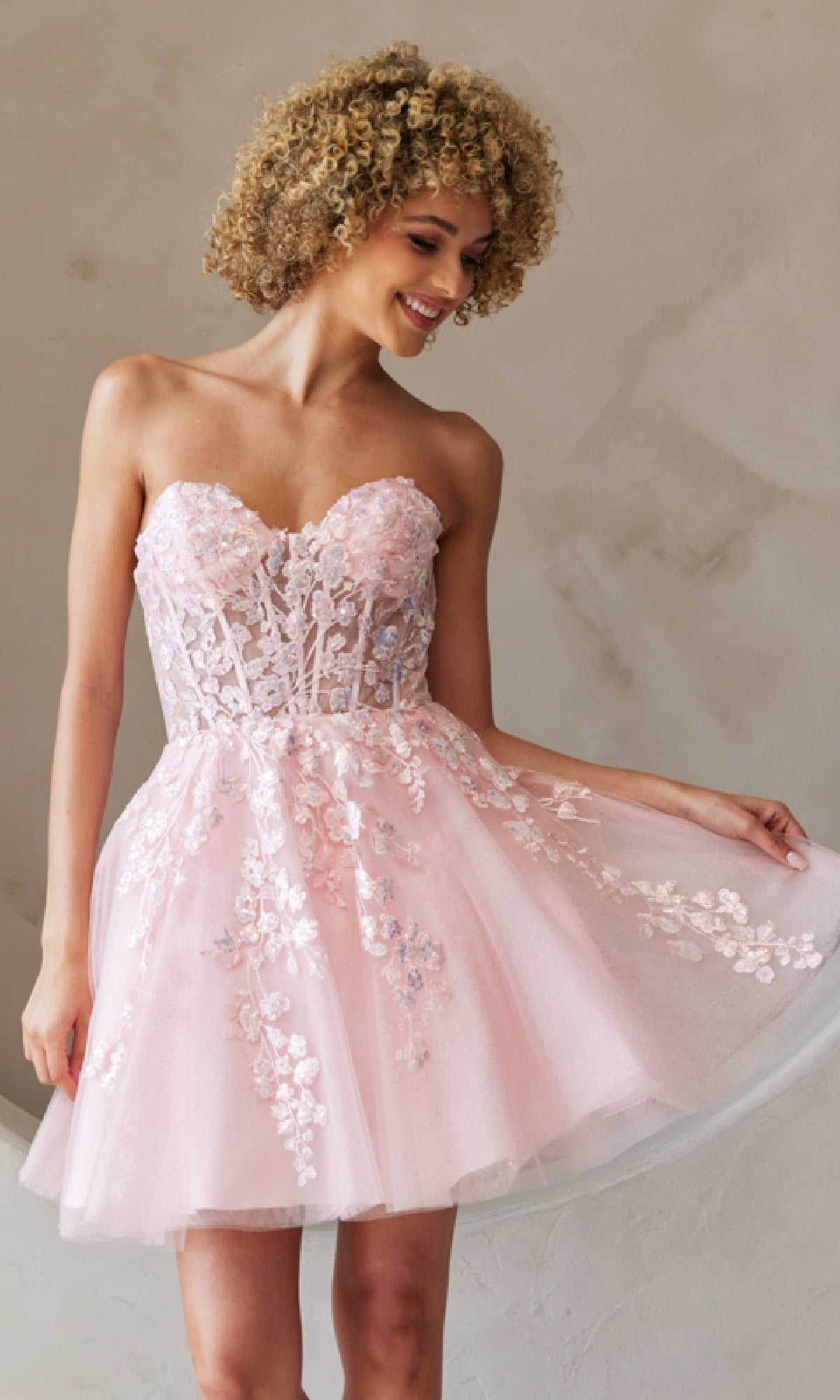 A woman poses by a light wall in the Strapless Short Pastel Homecoming Dress BZ9024S, adorned with light pink floral embroidery, holding out one side of her skirt.