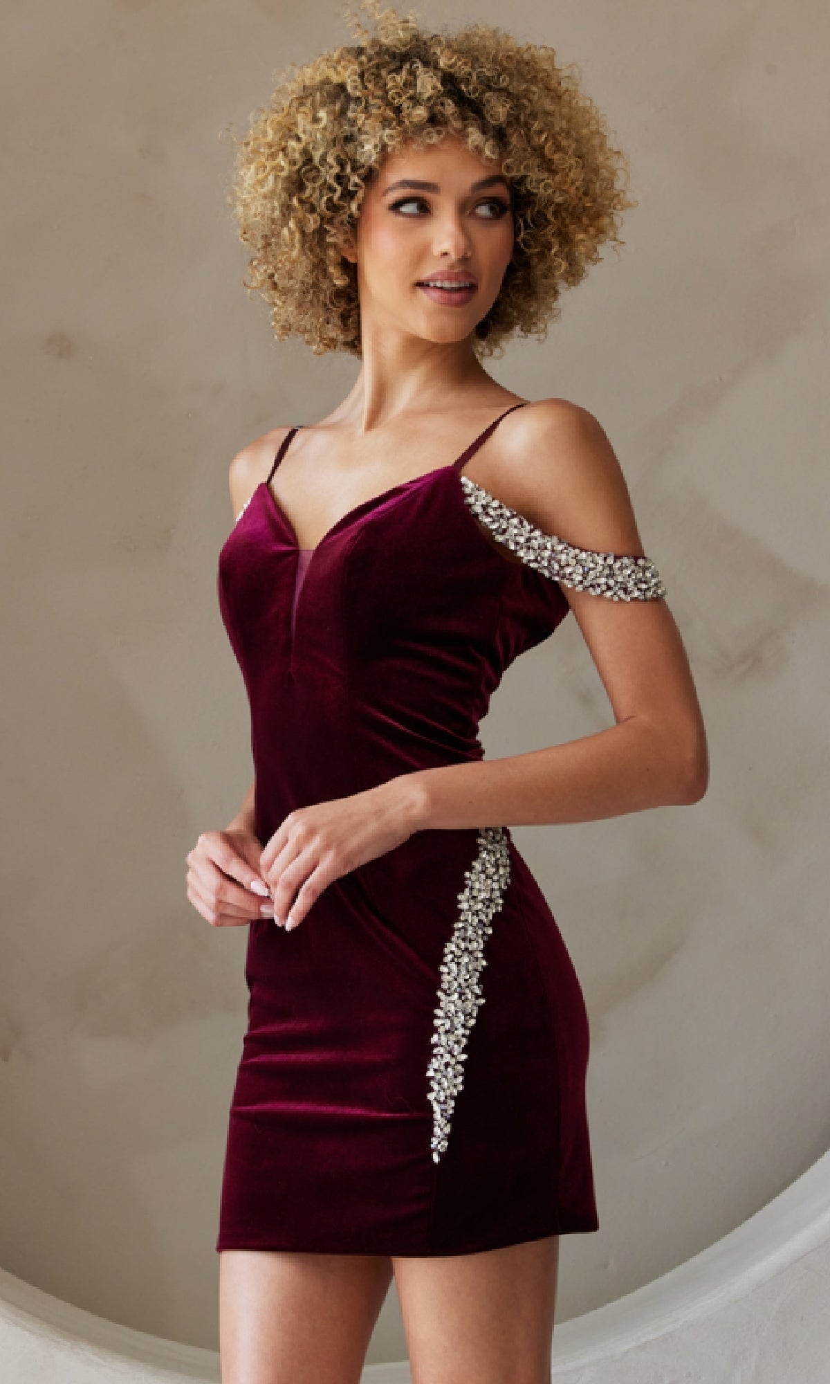 A woman wears the Bead-Trimmed Short Velvet Homecoming Dress 6200S, a fitted burgundy velvet dress with rhinestone accents on the strap and side, standing before a neutral background to showcase its elegant homecoming style.