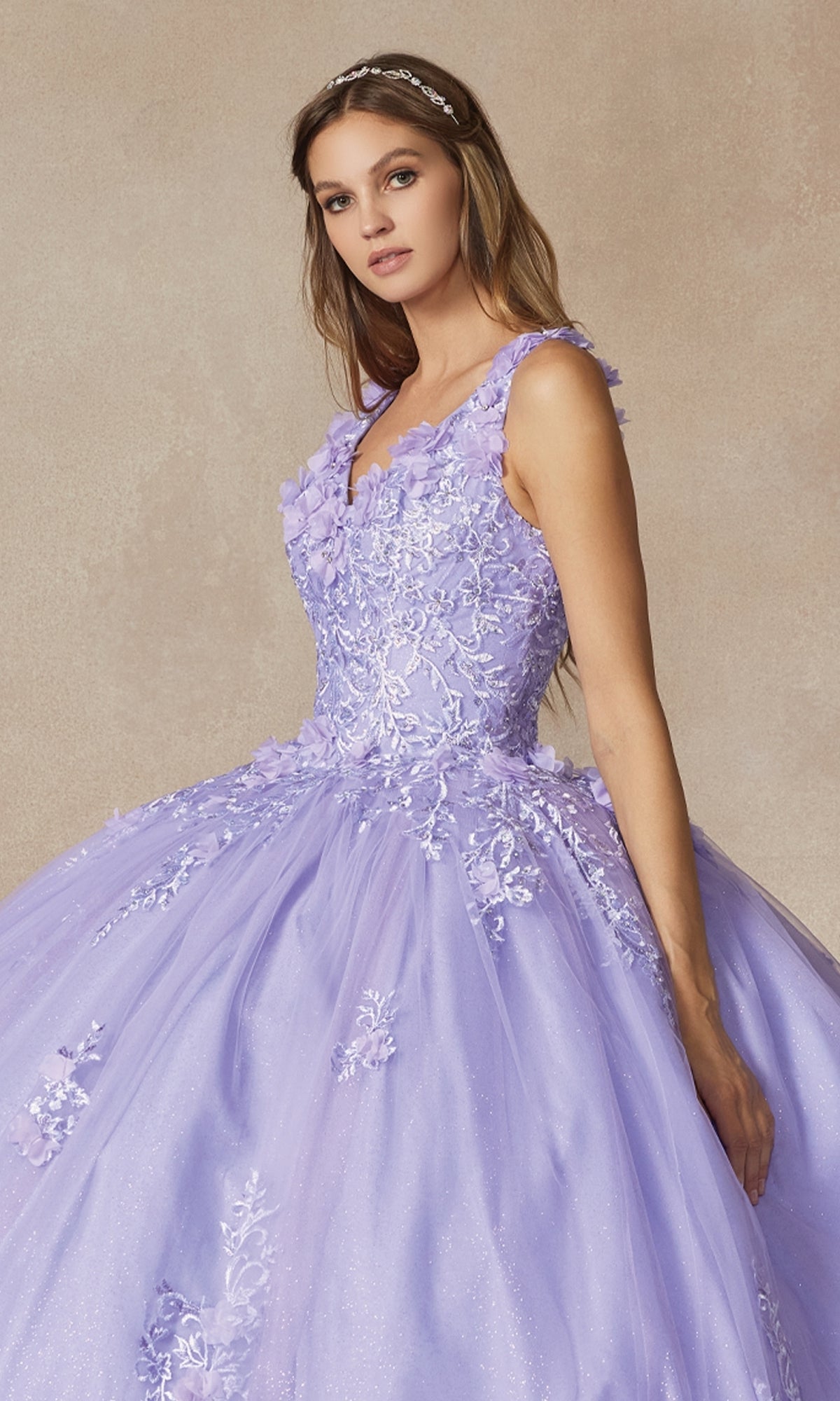 A woman wearing the Juliet 1437 Lilac Purple Quinceañera Dress with floral lace appliqués and a tiara stands against a neutral background.
