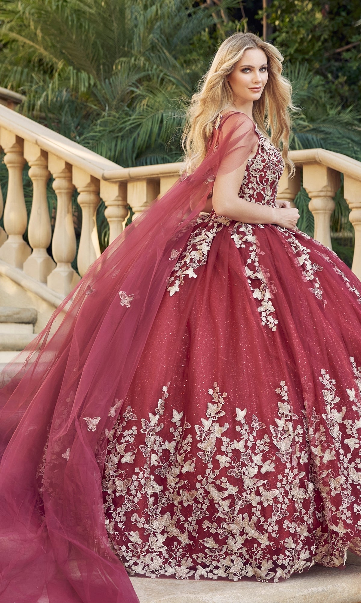 A woman stands on stone steps outdoors, surrounded by greenery, wearing the Juliet 1442 Quinceañera Dress with gold floral embroidery, deep red hue, and a long sheer cape.