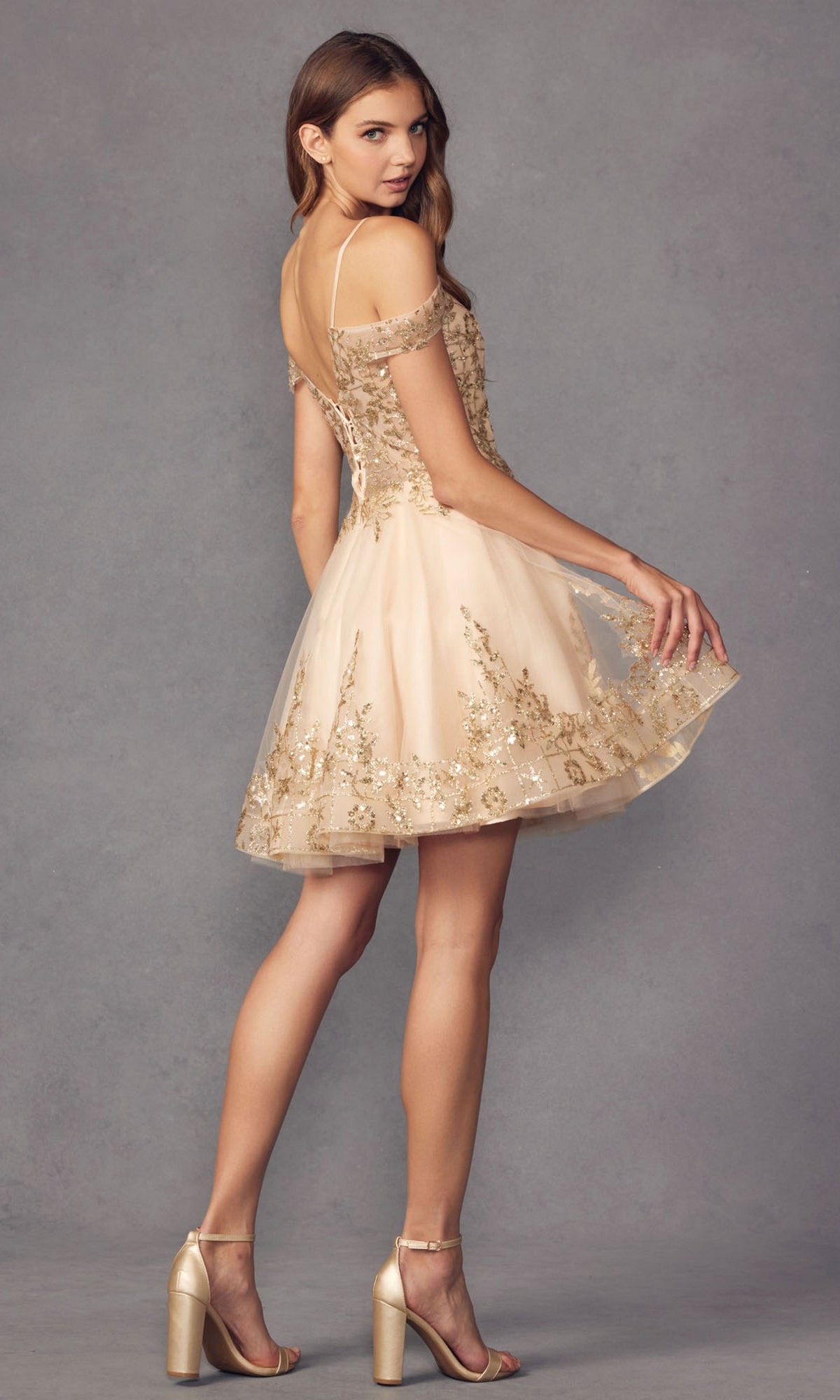 A woman in the Glitter-Print Short Babydoll Homecoming Dress 903, featuring a short off-shoulder beige design with gold embellishments and strappy heels, stands sideways against a gray background, exuding effortless elegance.