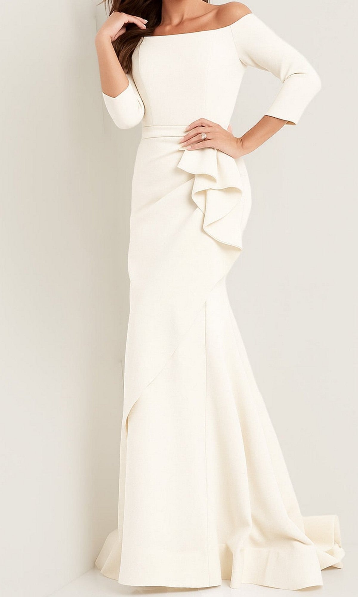 A woman stands against a light-colored wall wearing the Jovani Mother-of-the-Bride Long Formal Dress 00446 in ivory, featuring an off-shoulder neckline, long sleeves, and a floor-length skirt with ruffle detailing.