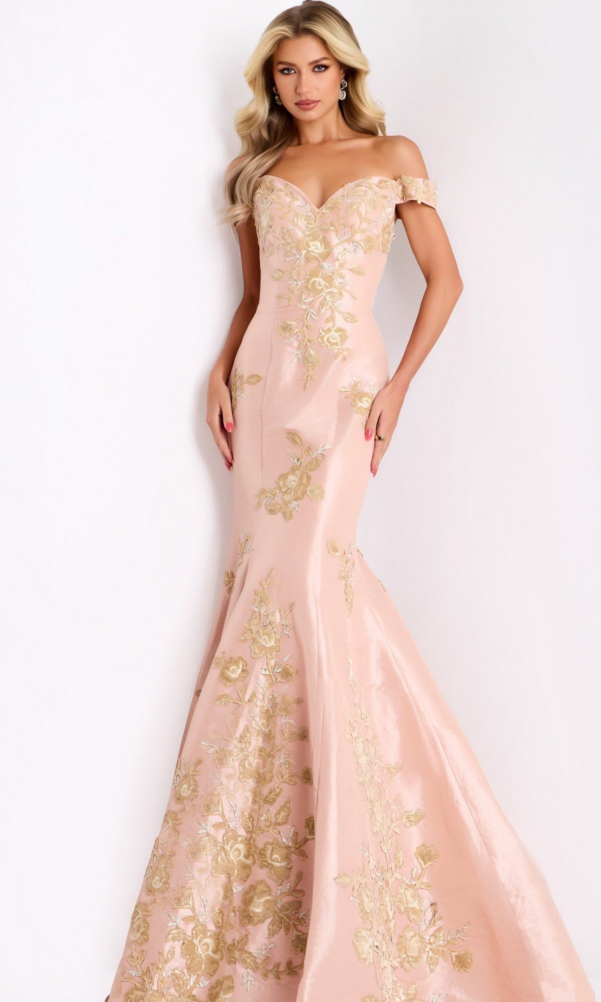 A woman wears the Jovani Gold and Black Long Prom Dress 00635 in blush and gold—a mermaid-style, off-the-shoulder gown adorned with gold floral embroidery—as she poses against a plain light background.