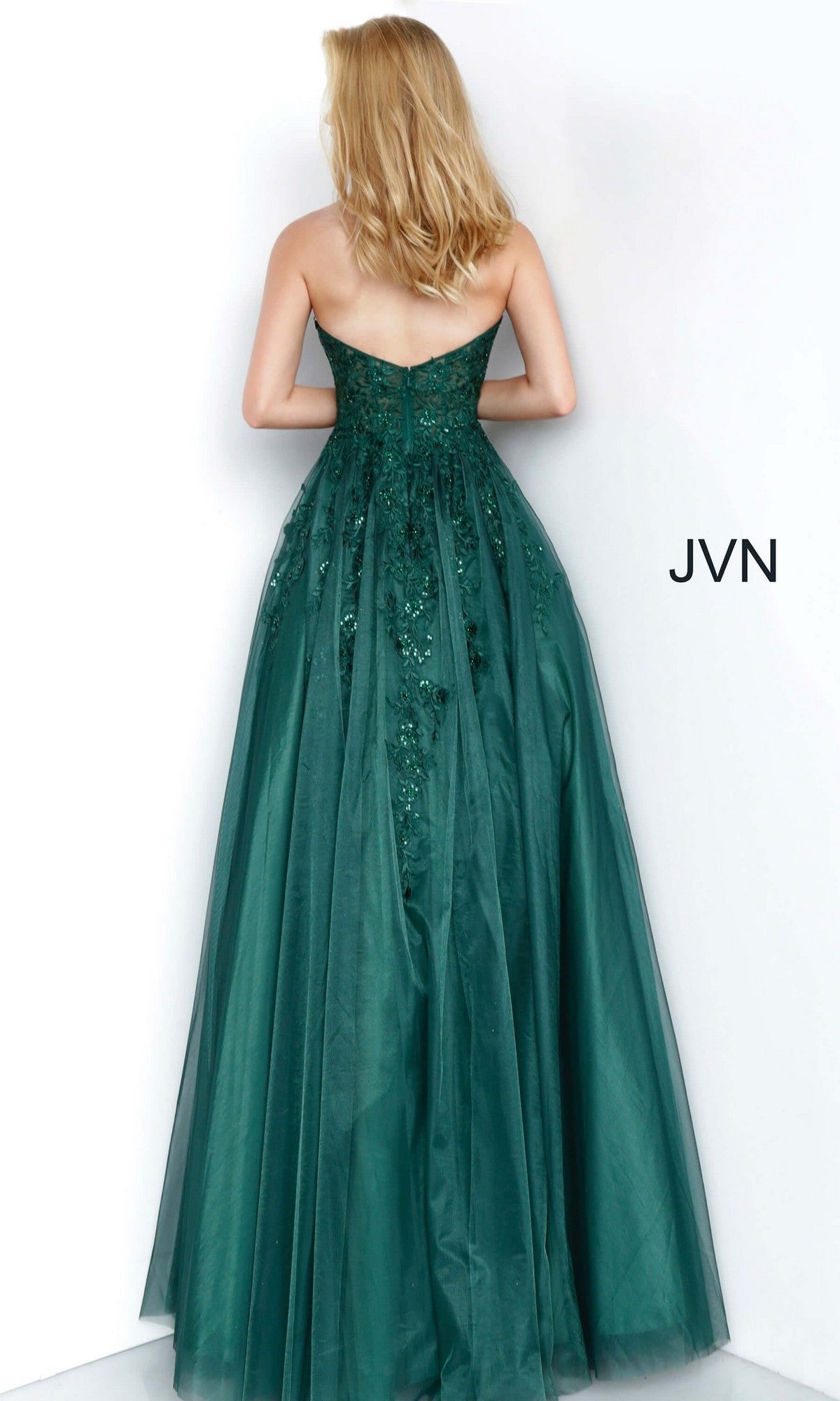 A woman wearing the Long Prom Dress: Jovani 00915, which features a strapless lace-embroidered bodice and a tulle ballgown skirt, stands facing a white wall with “JVN” printed to her right.