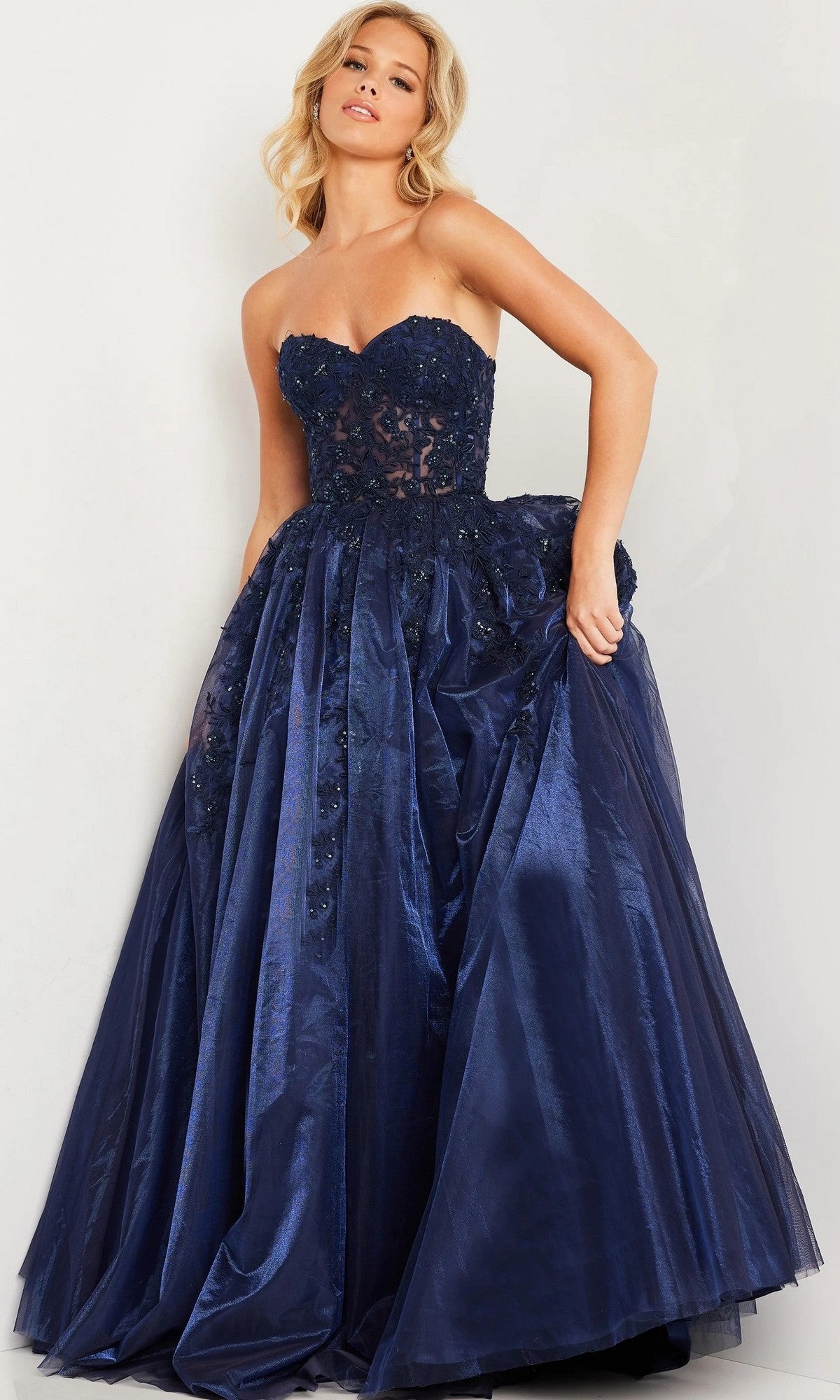 A woman wearing the Jovani 00915 Long Prom Dress in navy, which features a lace-embroidered, embellished fitted bodice and a shiny, voluminous tulle ballgown skirt, stands against a plain background.