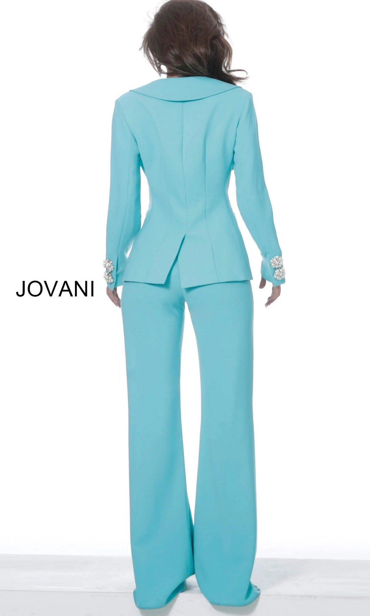 A person wearing the Jovani 02637 Long Jumpsuit in light blue with decorative cuffs stands with their back to the camera against a plain white background. The word “JOVANI” appears on the left side.