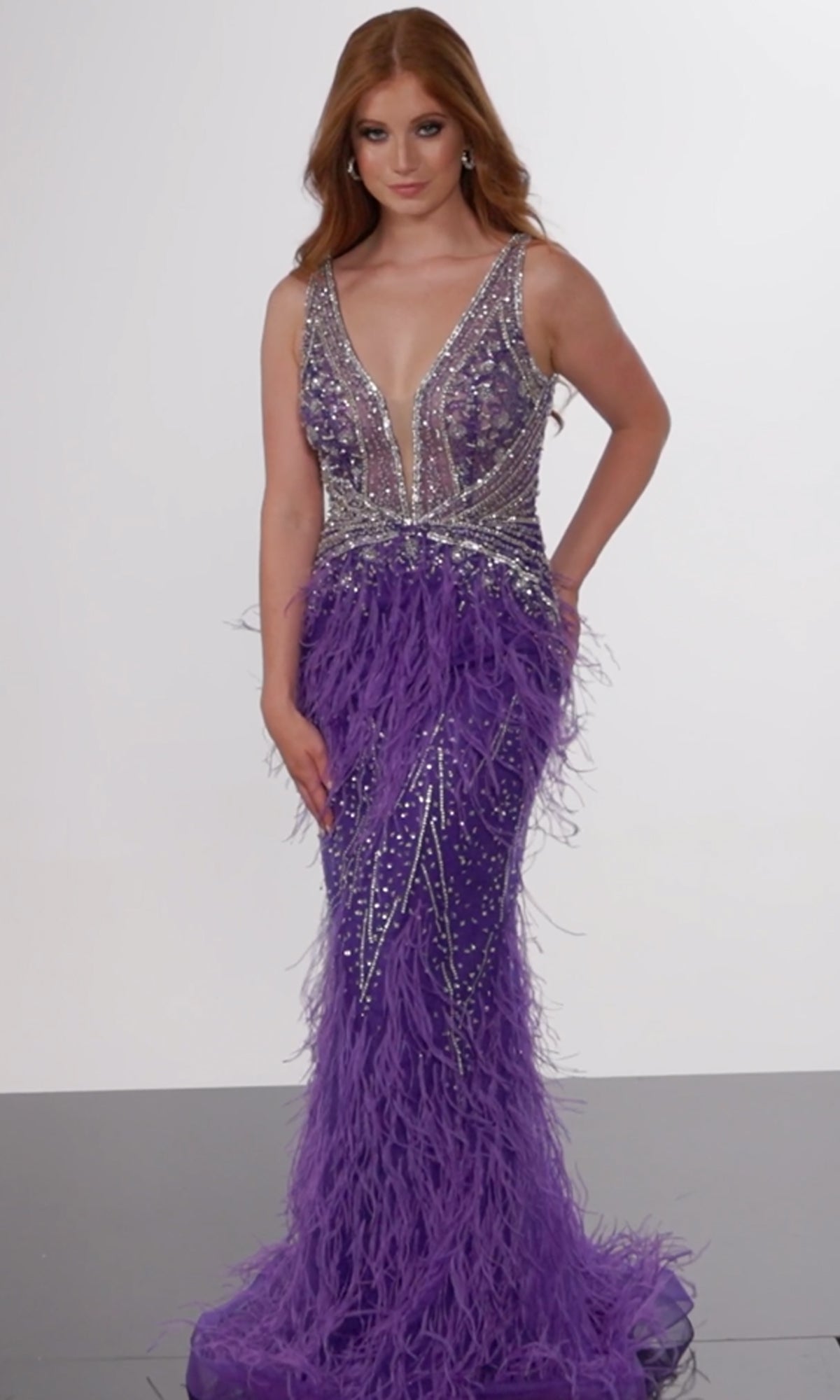 A woman wears the Jovani Sheer-Bodice Long Prom Dress with Feathers, featuring a sleeveless deep V-neck, silver beadwork, and a feather-embellished skirt, standing against a plain background.