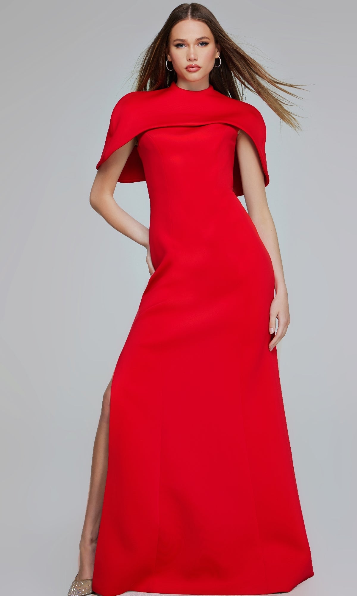 A woman poses confidently in the Jovani 06538 long formal dress, a floor-length bright red gown with an elegant cape detail, standing against a plain background.