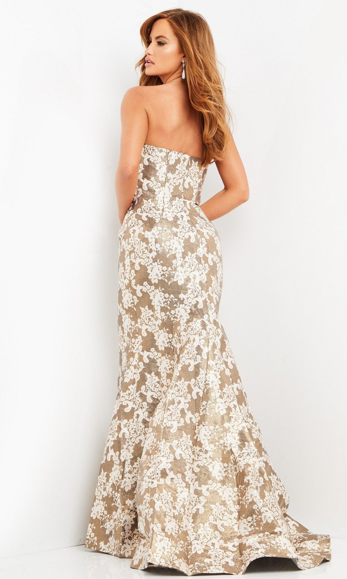A woman is seen from behind, wearing the Jovani 06760 Long Formal Dress—a strapless, floor-length jacquard gown featuring a tan base with a white lace floral pattern—while standing against a plain white background.