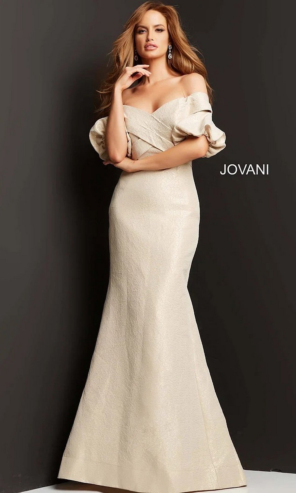 A woman poses against a dark background wearing the Jovani 06831 long formal dress—an off-the-shoulder, champagne-colored gown featuring puffed sleeves and a graceful mermaid silhouette.