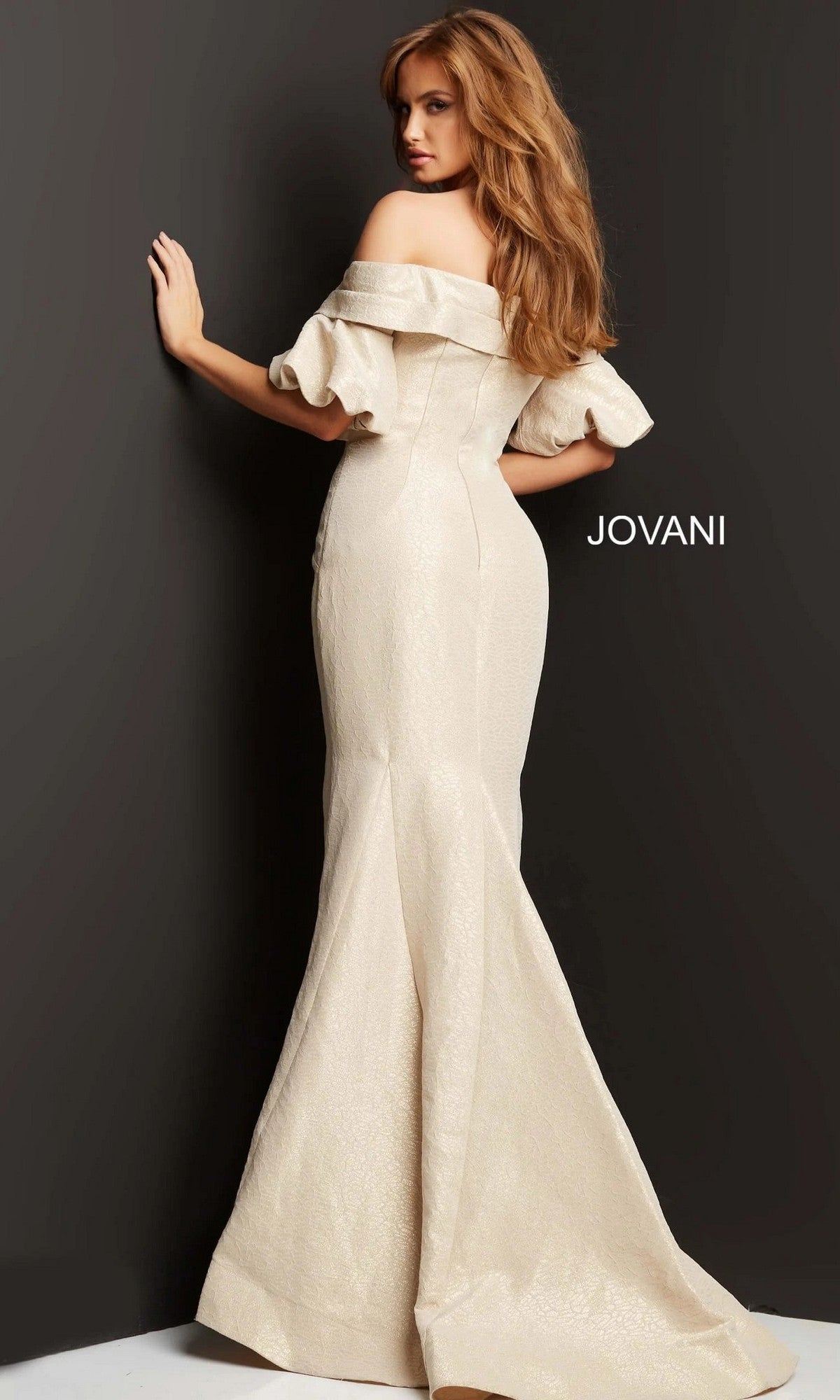 A woman stands against a dark wall with “JOVANI” written beside her, wearing the Jovani 06831 long formal dress—an off-the-shoulder, fitted gown in a cream-colored mermaid silhouette.