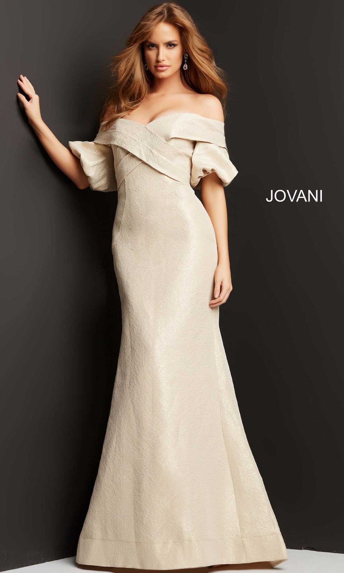 A woman stands against a dark wall wearing the Jovani 06831 long formal dress in champagne—a floor-length, off-the-shoulder gown with a fitted mermaid silhouette and textured fabric. The word “JOVANI” appears on the wall behind her.