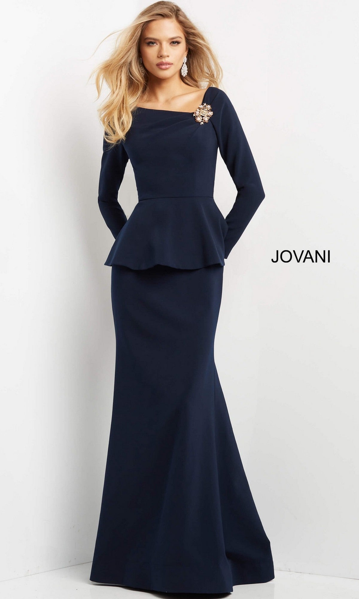 A woman wearing the Jovani 07131 long formal dress—a navy blue, long-sleeved peplum evening gown with a trumpet silhouette and a jeweled brooch at the shoulder—stands against a white background with “JOVANI” printed on the wall.