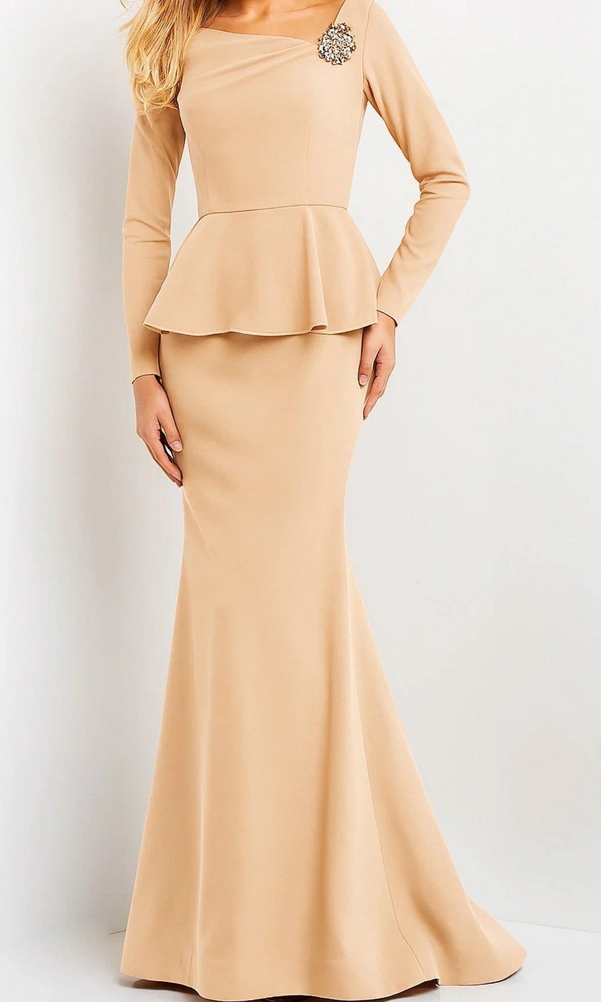 A woman wearing the Jovani 07131 long formal dress in champagne, featuring long sleeves, a peplum waist, and an elegant brooch detail on the shoulder, stands against a plain background.