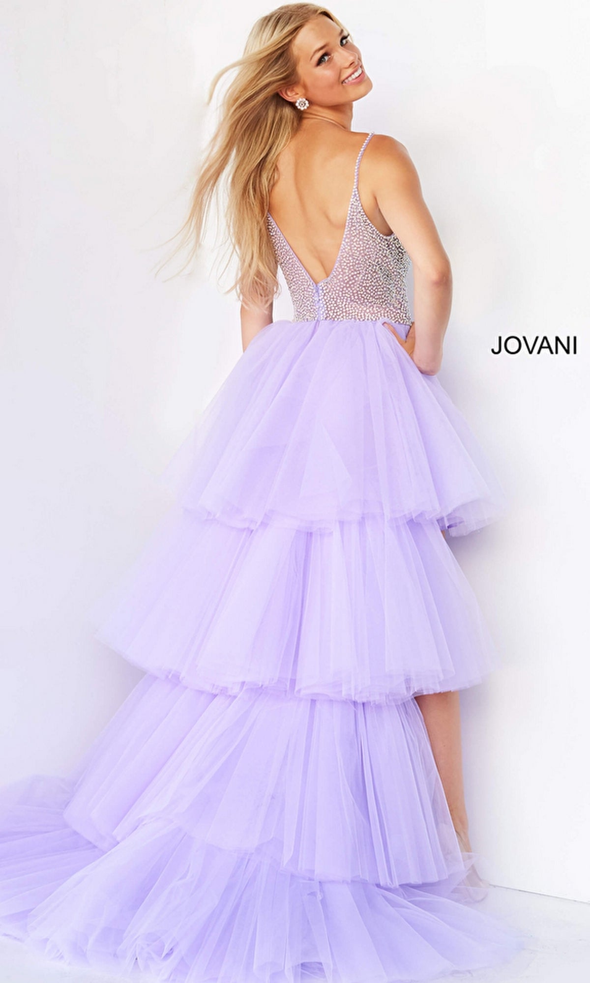 A woman smiles over her shoulder in a Jovani High-Low Sheer-Bodice Prom Dress 07231—lavender, sleeveless, backless, with an embellished bodice—posing against a white background.