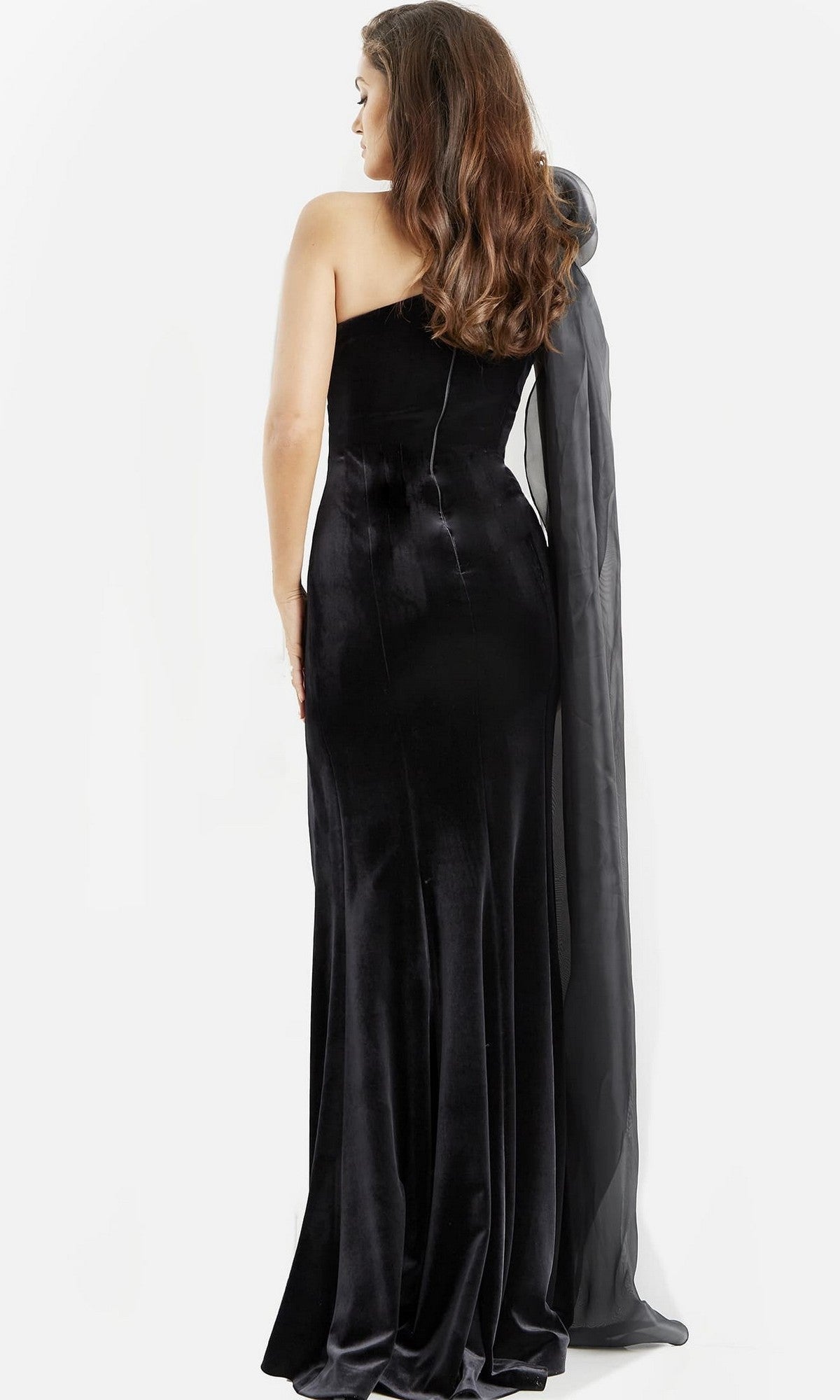 A woman in a black, floor-length Jovani 08116 one-shoulder velvet formal dress, accessorized with a long sheer black scarf, stands facing away against a white background.