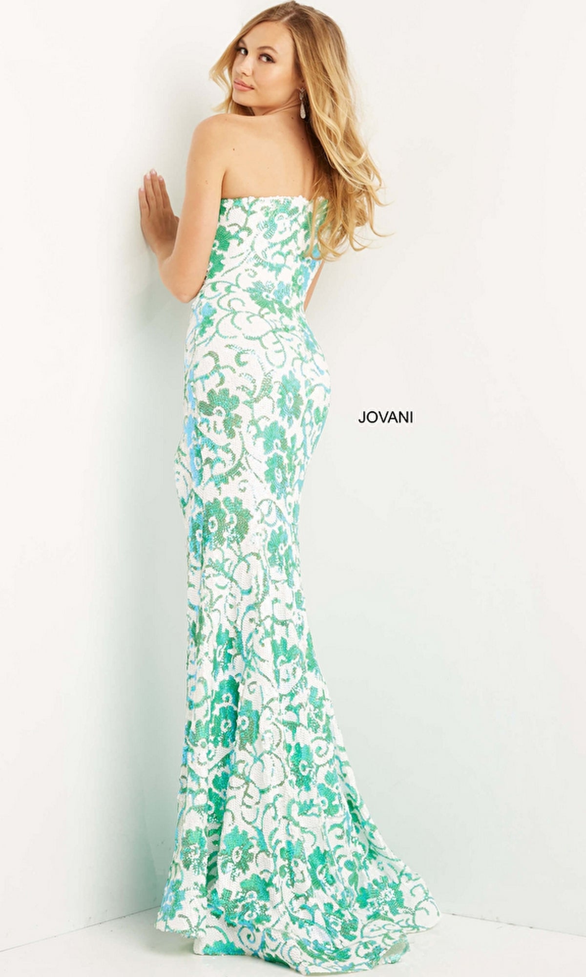 A woman poses against a white wall in the Jovani One-Shoulder Long Sequin Prom Dress 08256, a floor-length designer gown featuring elegant white and green floral patterns.