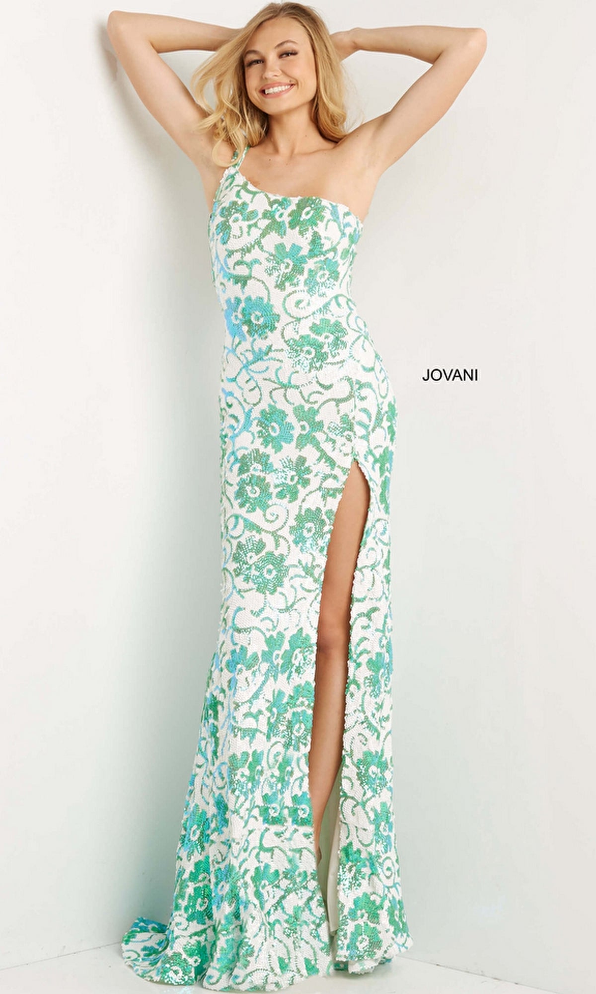 A woman poses with arms raised, smiling in the Jovani One-Shoulder Long Sequin Prom Dress 08256 in Ivory/Emerald, featuring a fitted silhouette and high slit, against a white wall.