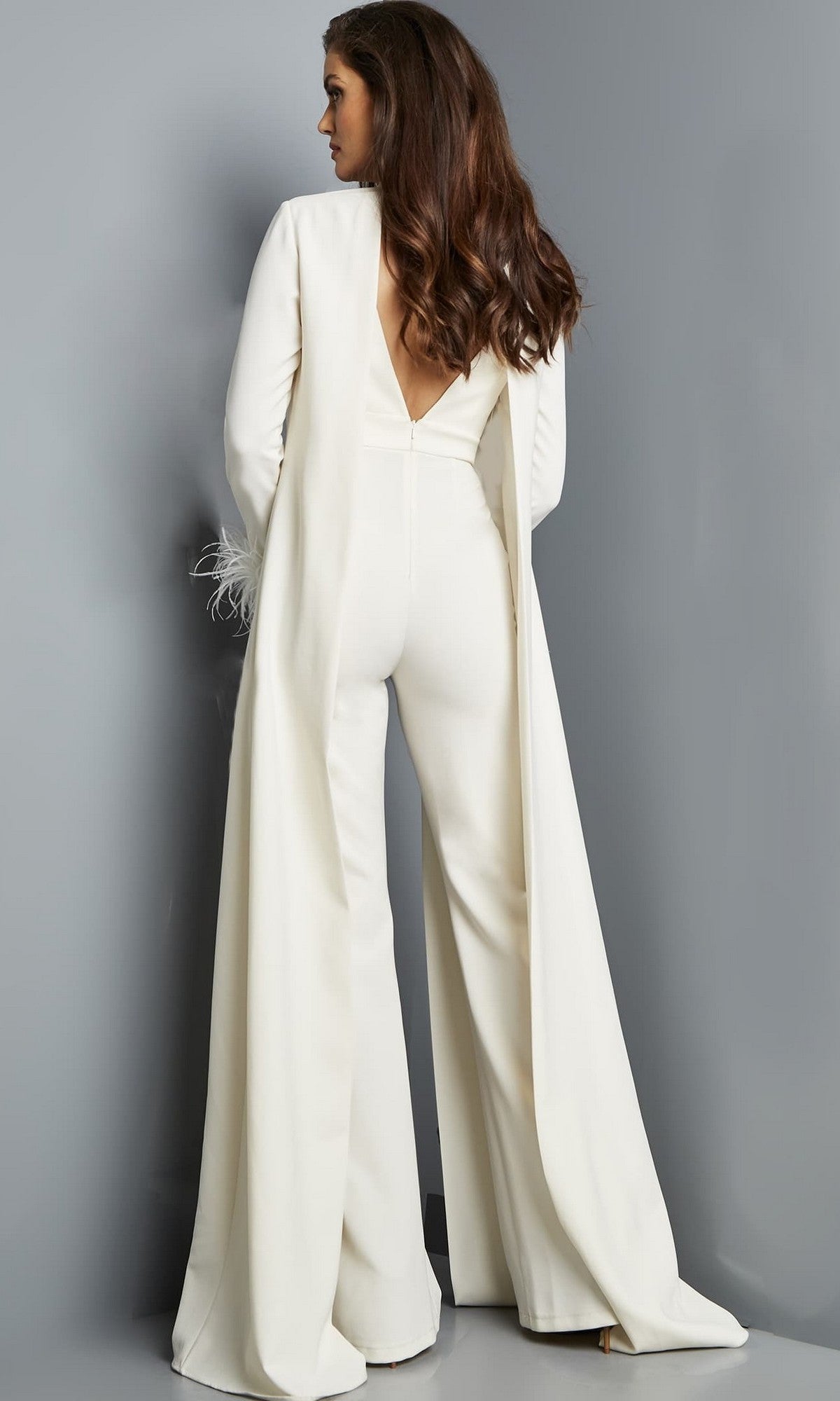 A woman stands facing away against a gray background, wearing the Jovani 09479 long jumpsuit in ivory crepe, featuring sleeveless styling, wide legs, and a dramatic cape-like overlay.