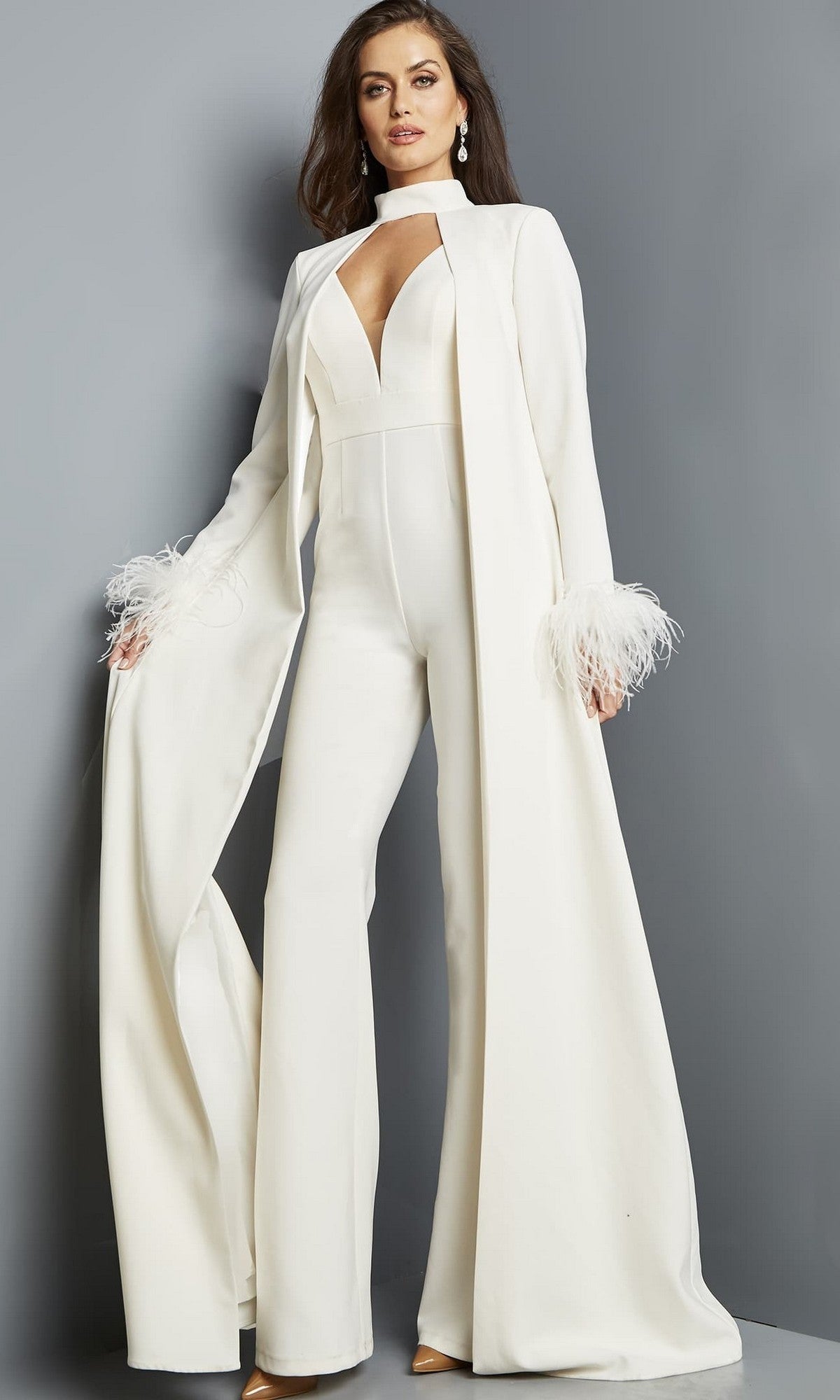An elegant woman wears the ivory Long Jumpsuit: Jovani 09479, featuring a formal deep V-neckline and a long matching cape with feathered cuffs, standing against a plain gray background.