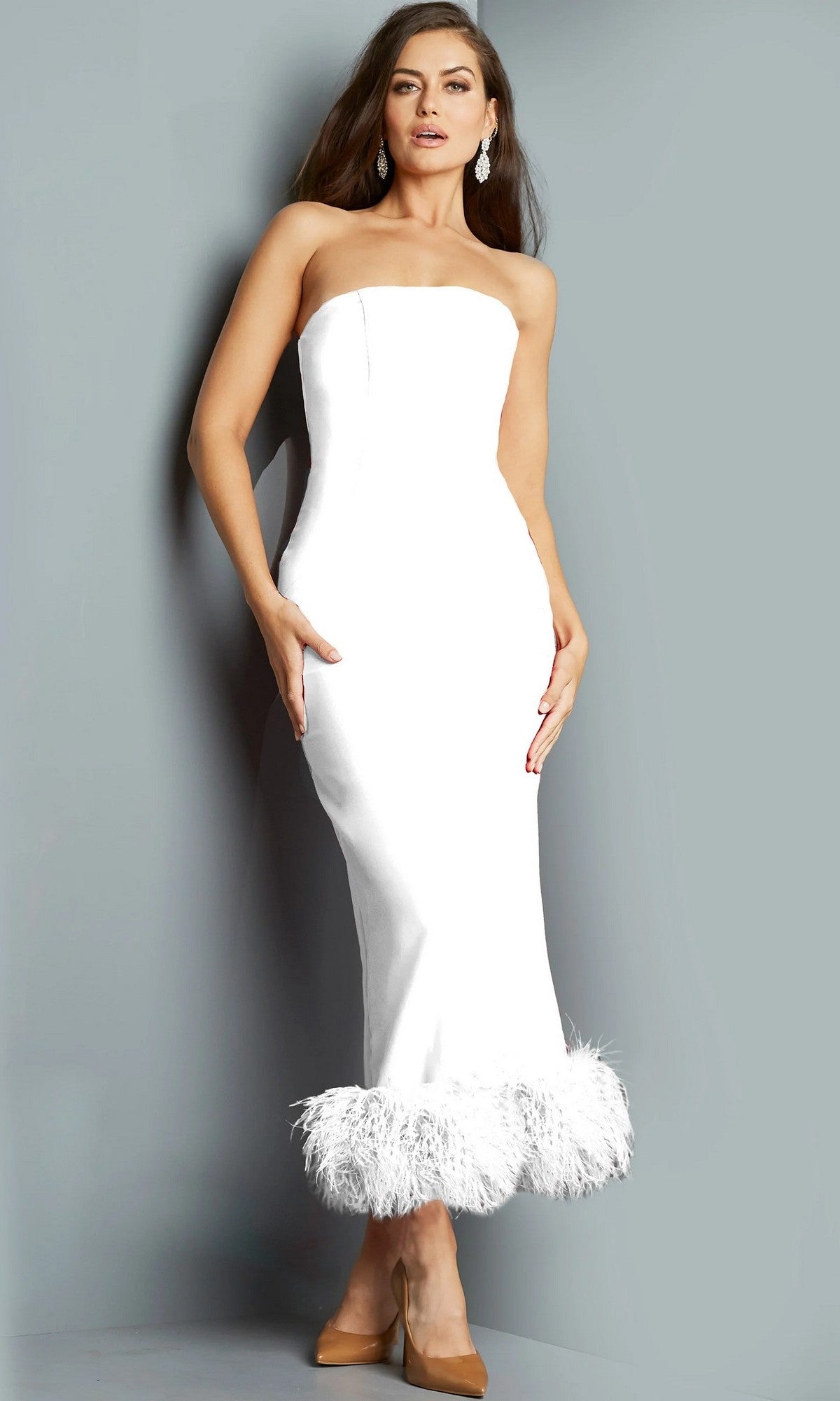 A woman wearing the white Jovani 09630 long formal dress, which is strapless with a feathered hemline, styled with tan pointed shoes and statement earrings, stands against a plain gray background.