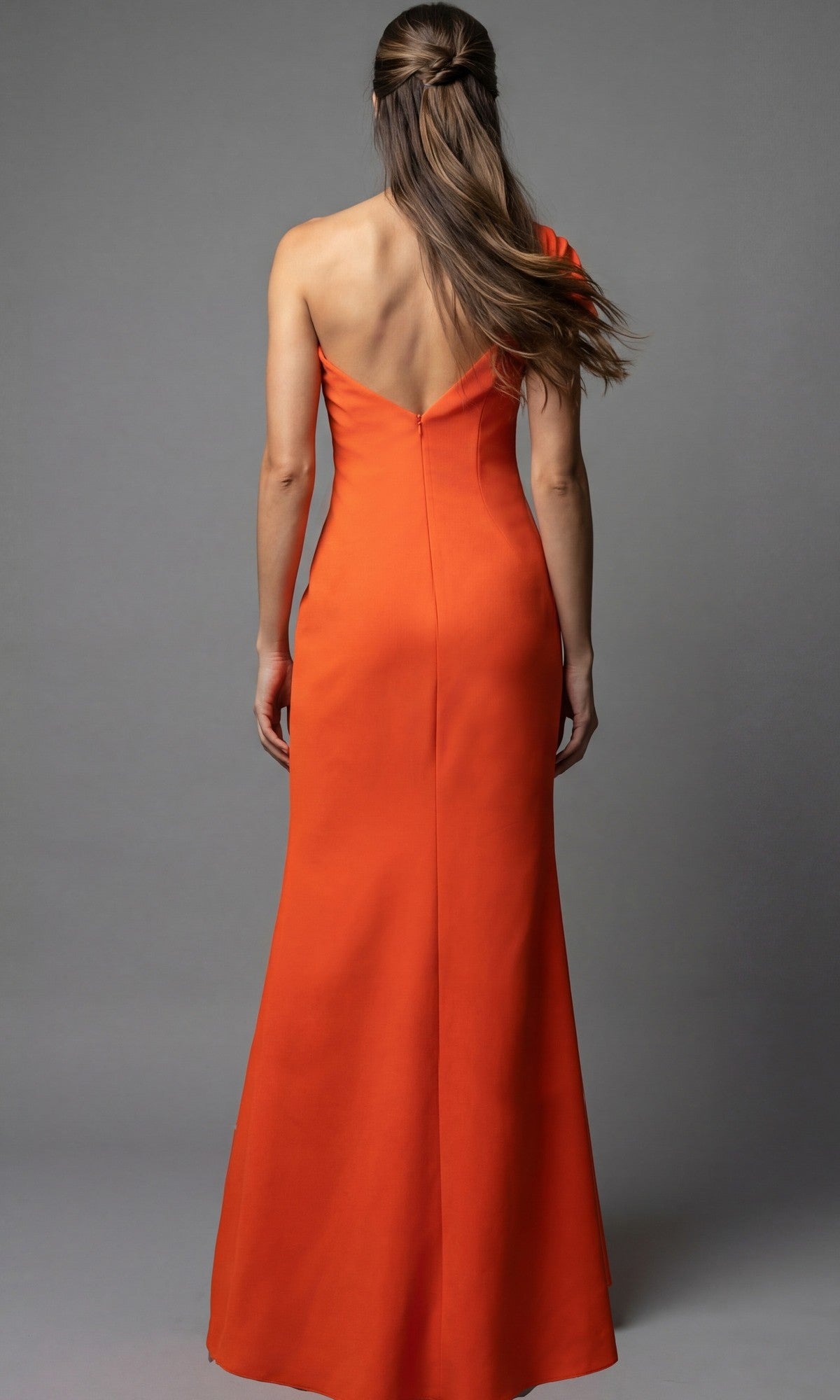 A woman wearing the Jovani 09961 long formal dress in orange stands facing away, showcasing its V-shaped back and one-shoulder design, with straight hair against a plain gray background.