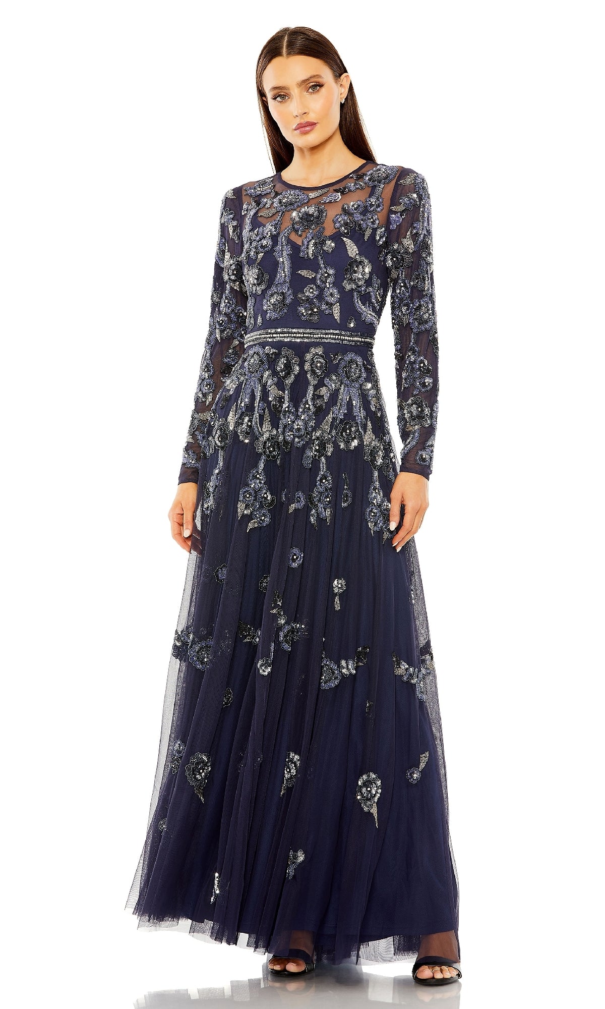 A woman stands against a white background wearing the Mac Duggal 10013 long sleeve blue formal dress featuring silver floral embroidery and a sheer neckline.