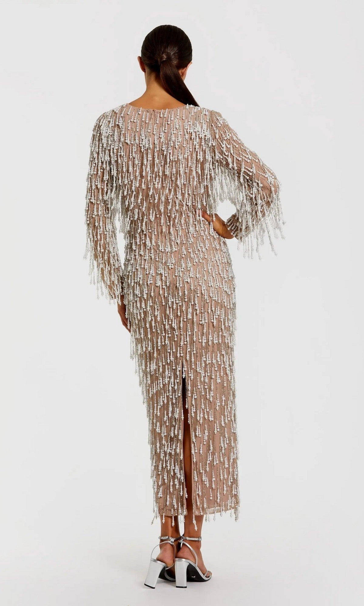 A woman with her hair in a low ponytail faces away, wearing the Midi Wedding Guest Dress: Mac Duggal 10120— a sheer nude midi dress with pearl beaded fringes—paired with metallic high-heeled sandals.