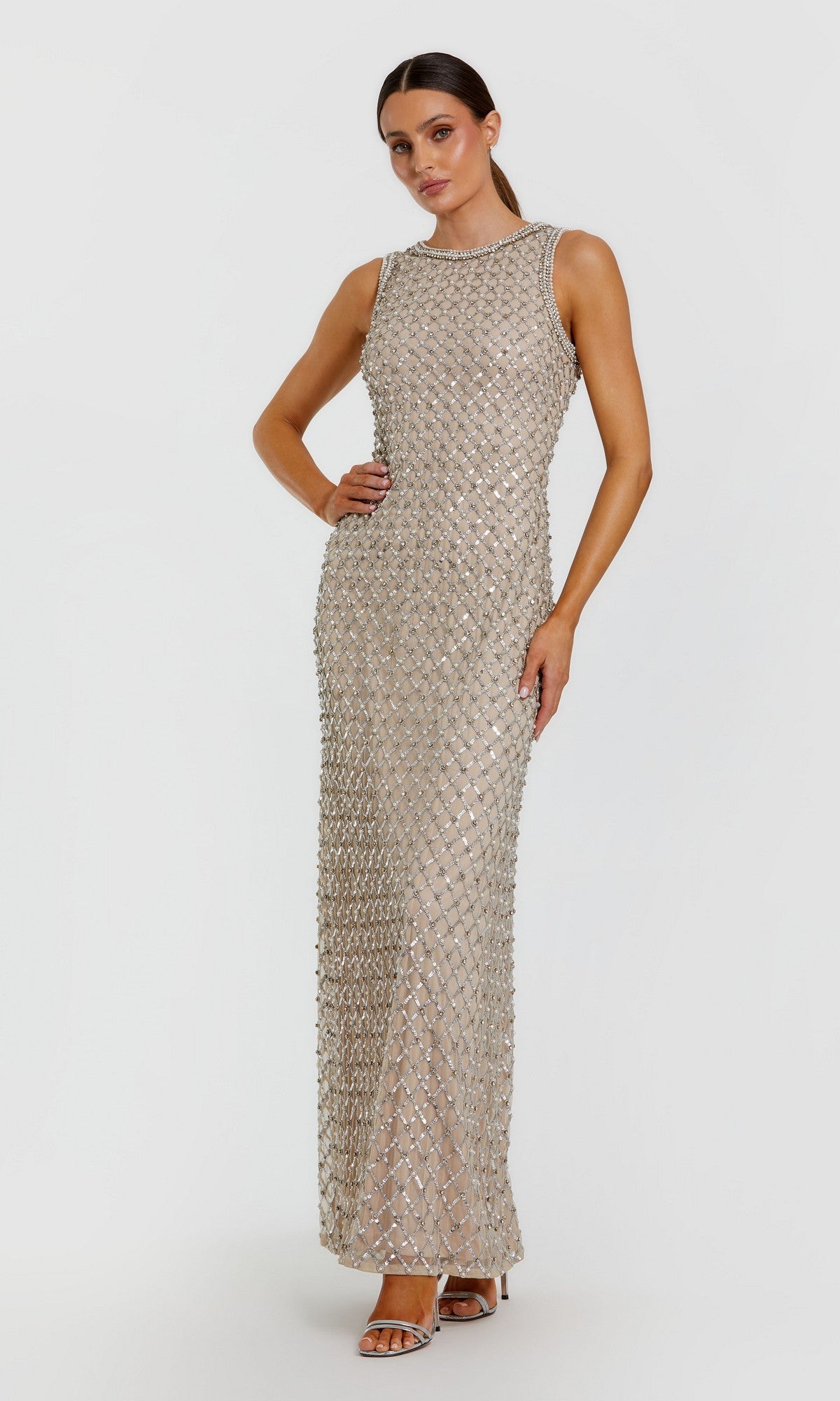 A woman stands against a plain white background wearing the Mac Duggal 10129 long formal dress in Silver Nude—a sleeveless, floor-length gown adorned with silver sequins and featuring a high neckline. She completes the look with open-toe heels.