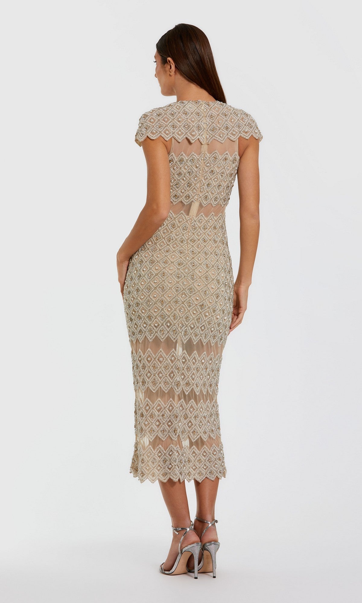 A woman wearing the Long Formal Dress: Mac Duggal 10147—a short-sleeve, knee-length beige sheath with geometric beadwork—stands in profile with her back partially turned, paired with silver high-heeled sandals against a plain background.