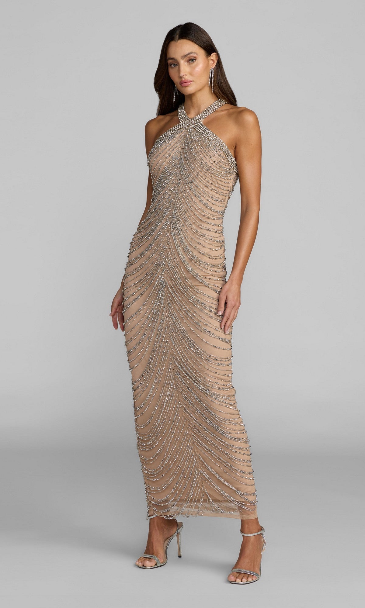 A woman wearing the Mac Duggal 10206 sleeveless long formal dress in nude silver, featuring draped beadwork and silver beaded detailing, stands in high-heeled sandals against a plain light gray background.