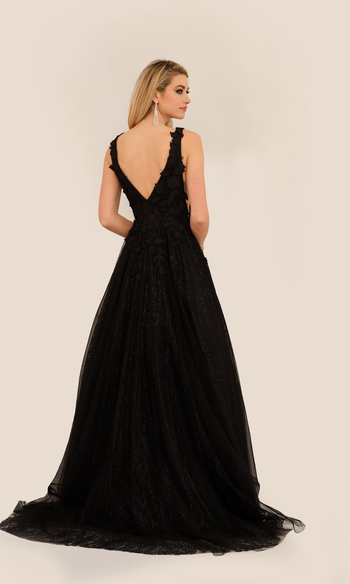 A woman wearing the Long Formal Dress A10288 by Dave and Johnny, a sleeveless, floor-length black ball gown with a deep V-back, stands and looks over her shoulder against a neutral background.